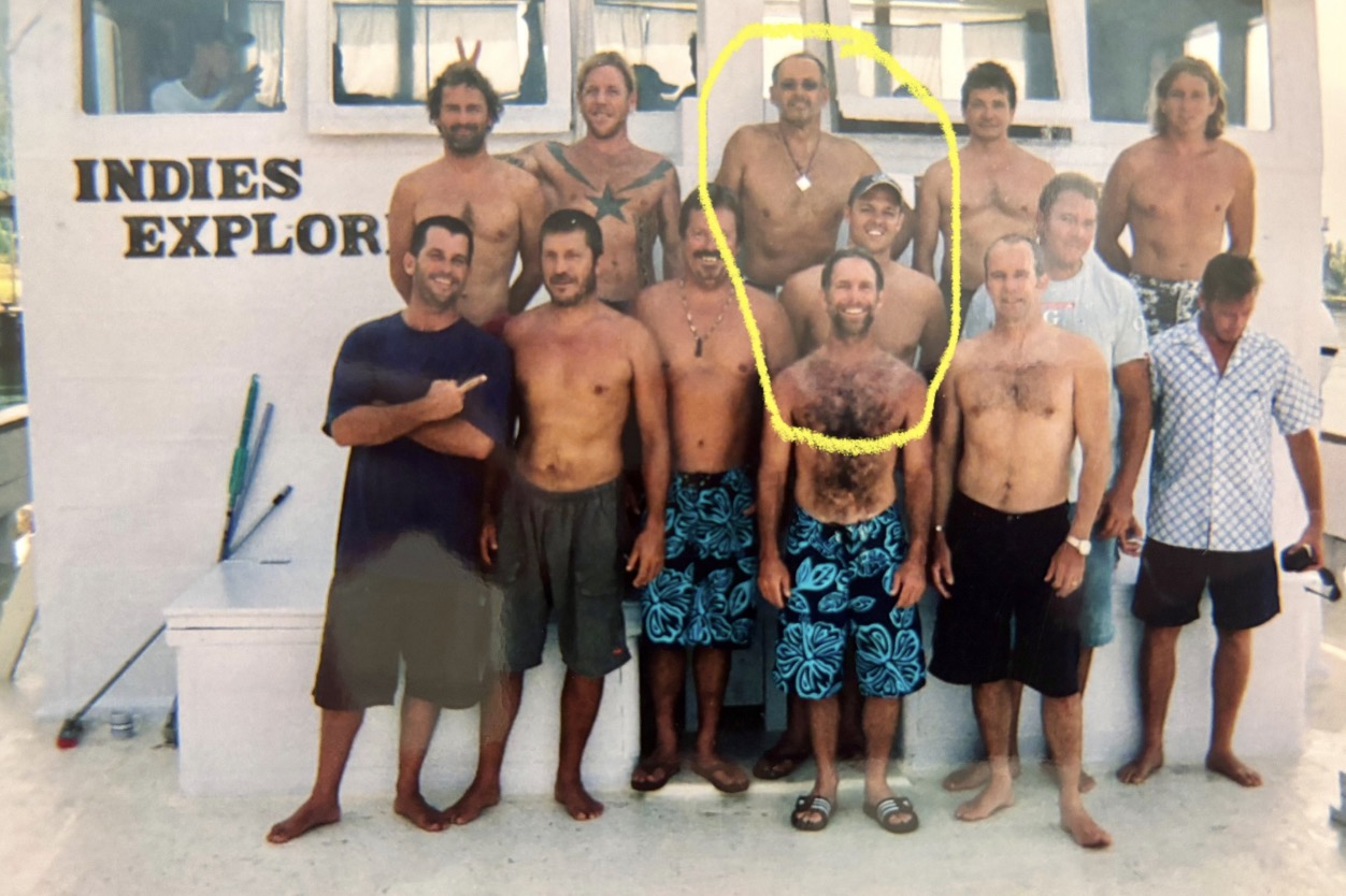 Group of men in swimwear and casual clothing on a boat deck, with a sign reading 'Indies Explorer' in the background.