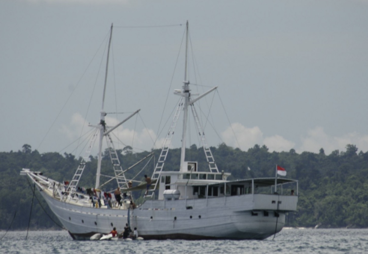 Large white boat with two masts on water, with a forested shoreline in background.