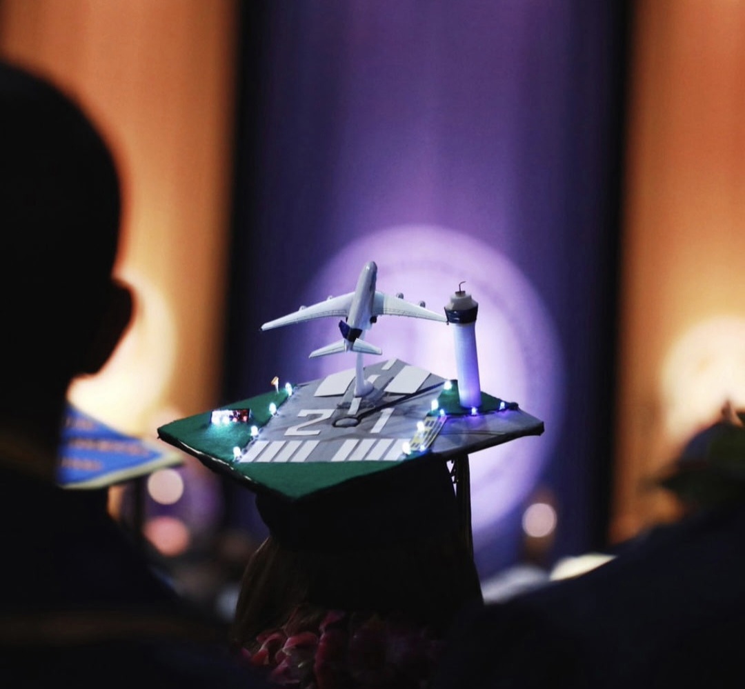 Person holding a graduation cap with a miniature airplane and airport runway model, audience in background, stage lighting.