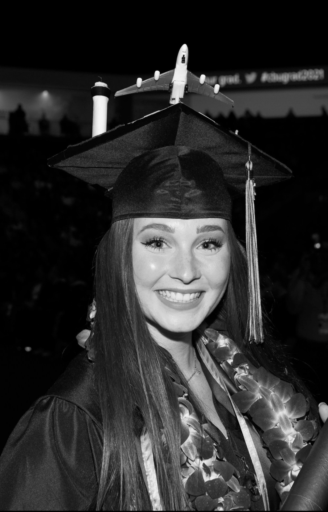 Young woman in graduation cap and gown smiling at graduation ceremony, background dark, visible stage lights.