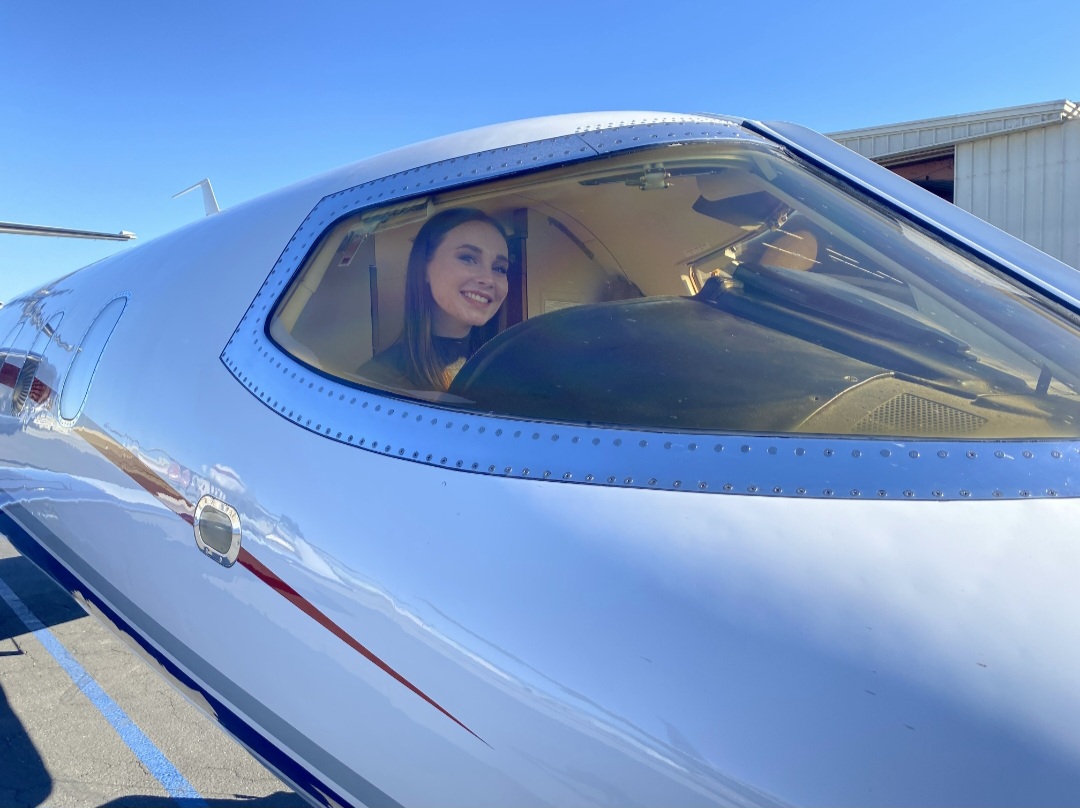 Woman smiling inside an airplane cockpit, viewed through the windshield, with a clear blue sky in the background.
