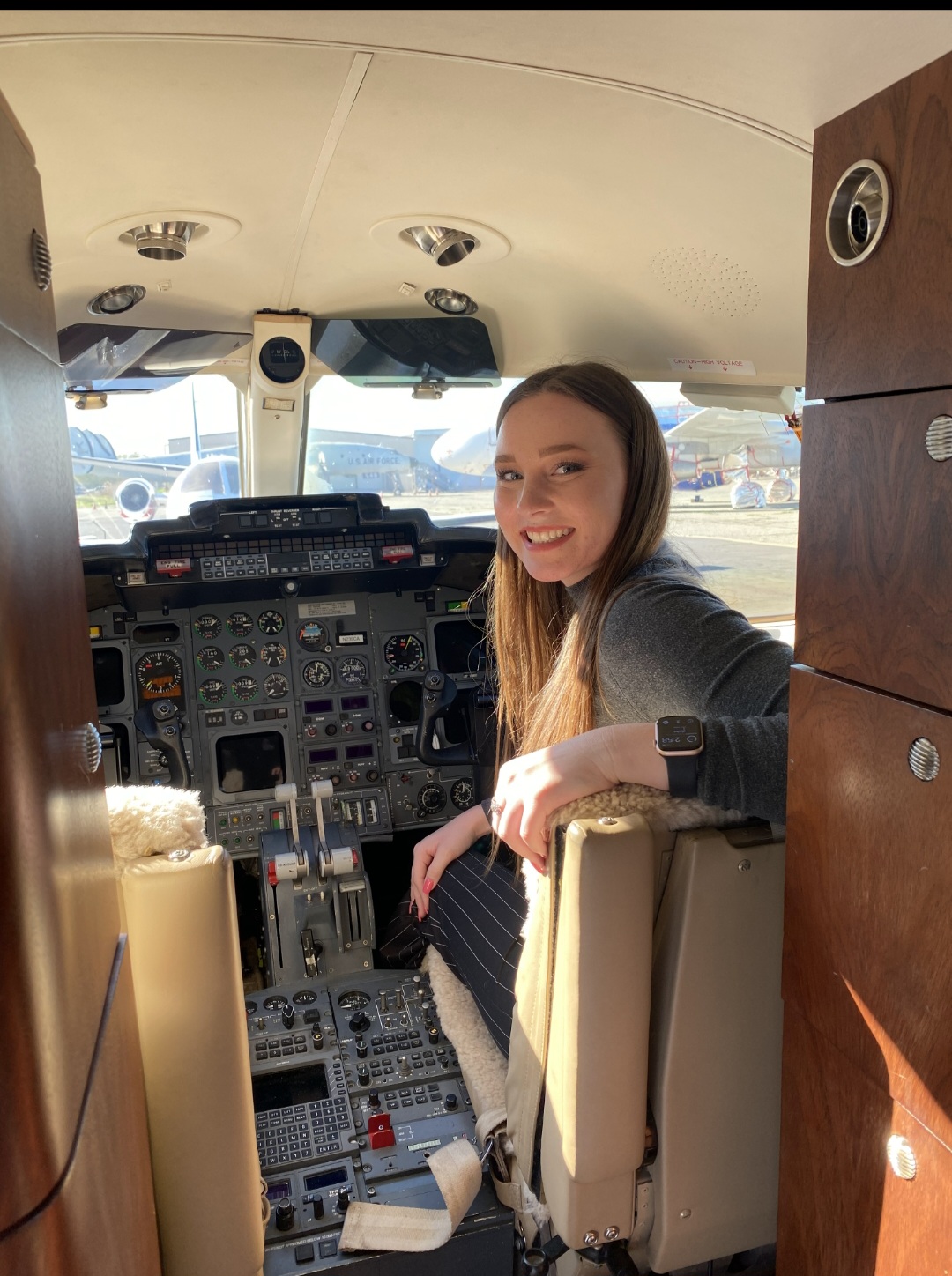 Young woman sitting in cockpit with aircraft controls, smiling, with windows showing airport outside, sunlight illuminating interior.