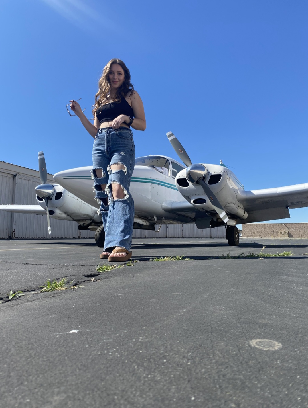 Young woman with long hair in ripped jeans and a black top stands in front of a small airplane on tarmac under a clear blue sky.