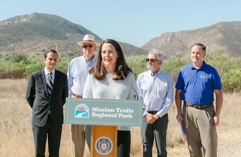 Group of five people standing outdoors with mountains in background, one woman speaking at a podium, smiling.