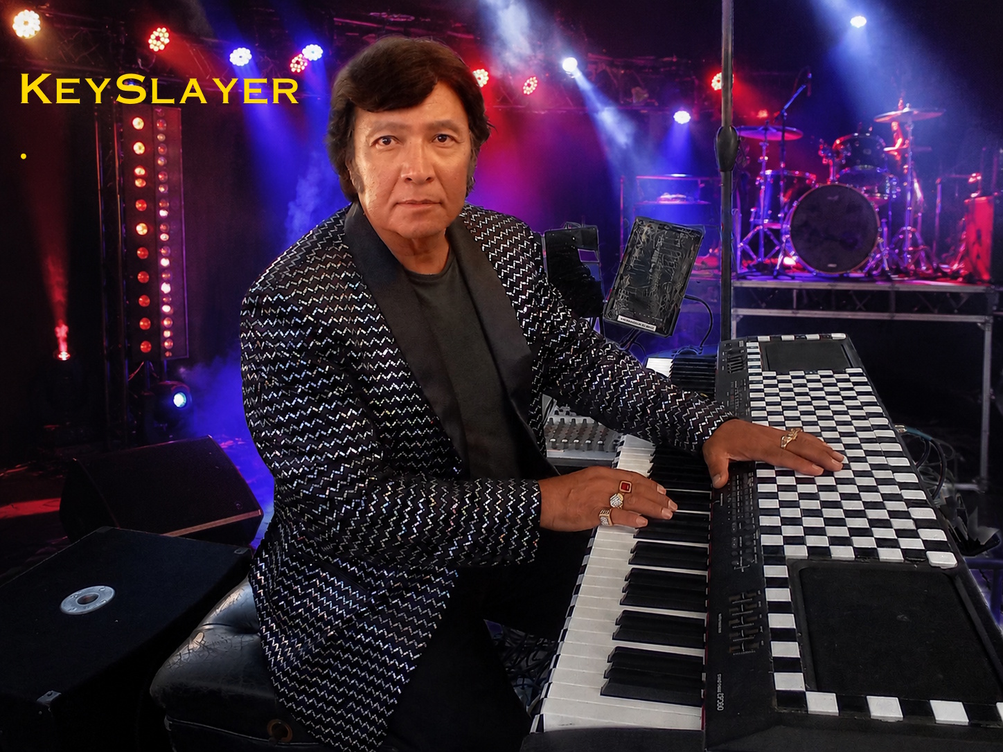 Man playing keyboard on stage with colorful lights and drums in background, wearing a patterned blazer.