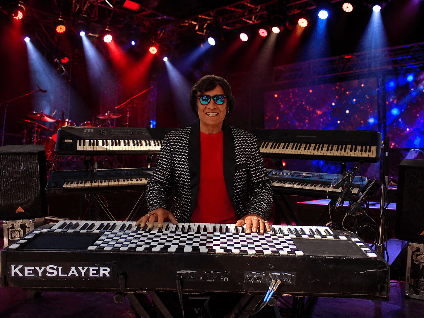 Person playing a keyboard on stage with colorful lights and screens in the background.