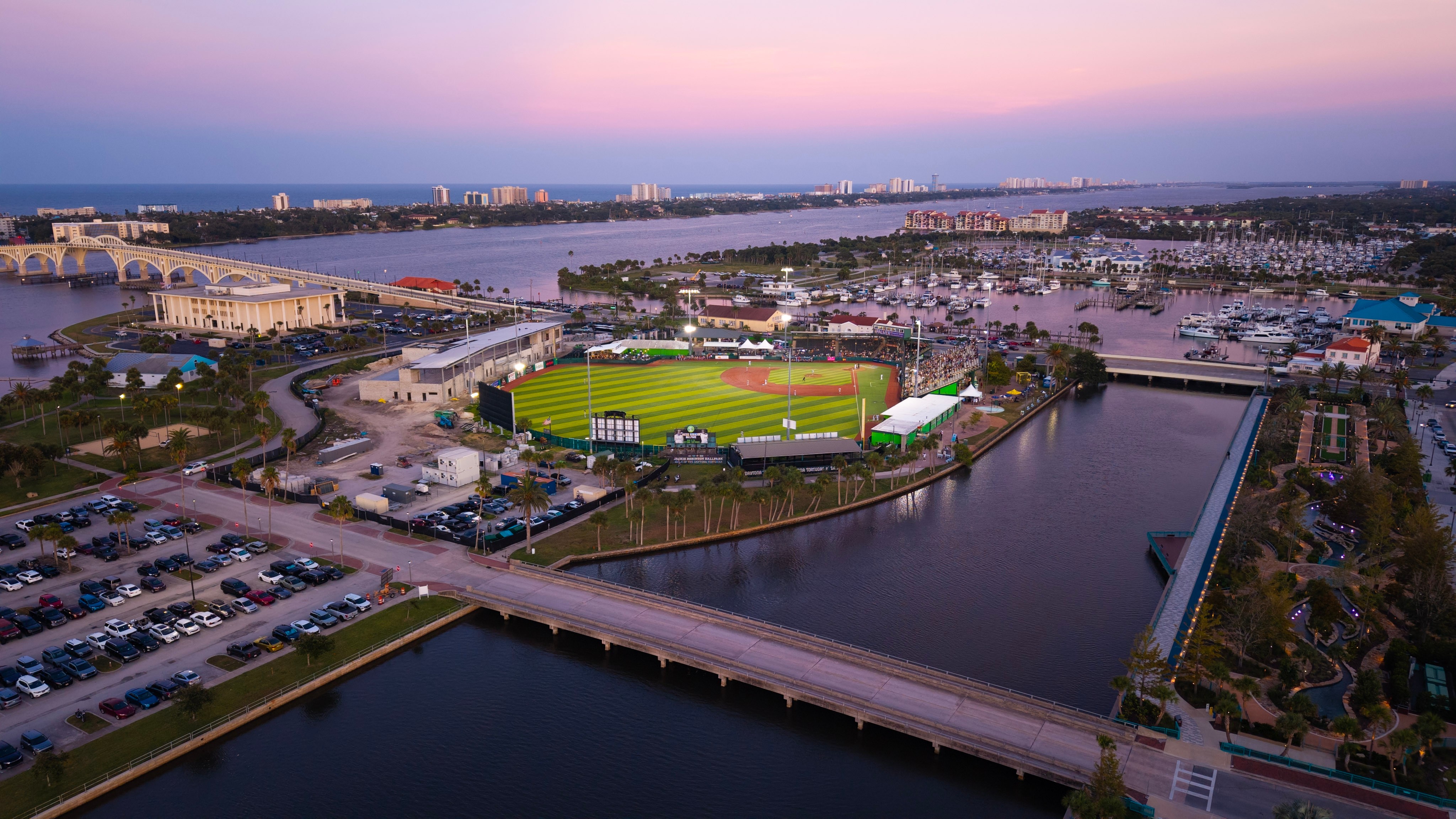 Aerial view of a sports stadium, parking lot, waterway, and cityscape at sunset.