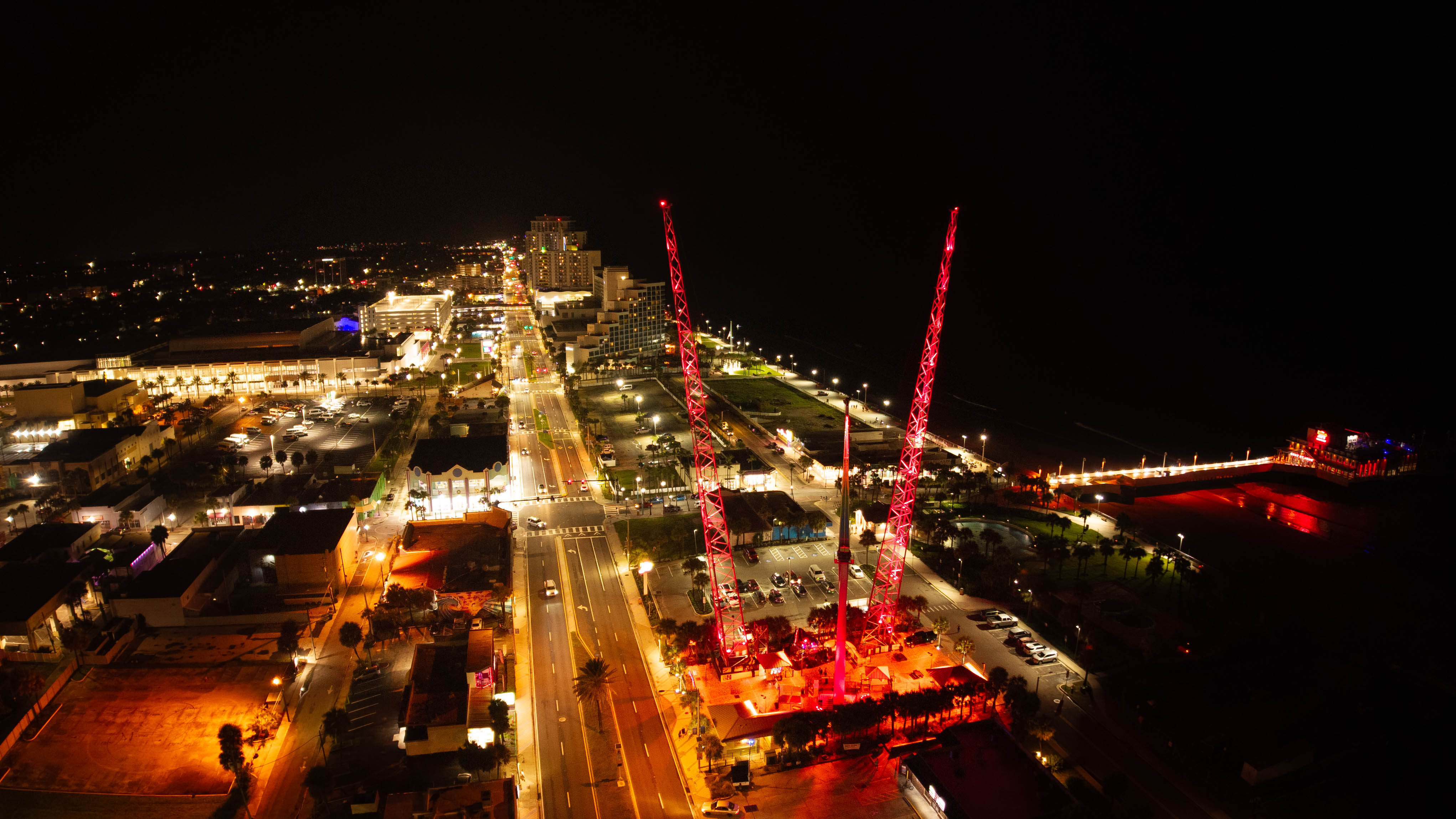 Night cityscape with illuminated streets and two tall red-lit structures, possibly cranes, against a dark sky.