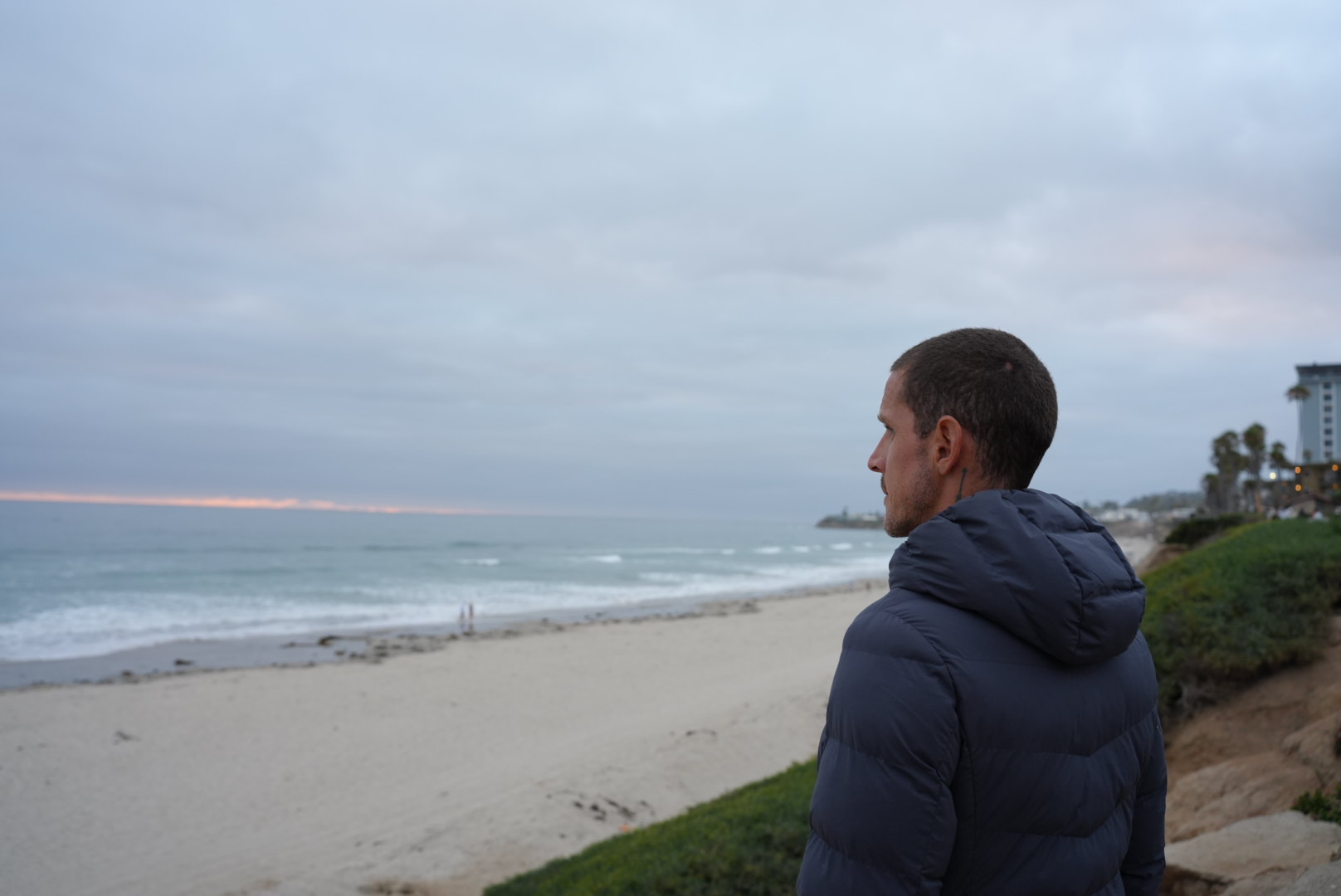 Man in a dark jacket looking at the beach and ocean with cloudy sky, shoreline, and distant buildings.