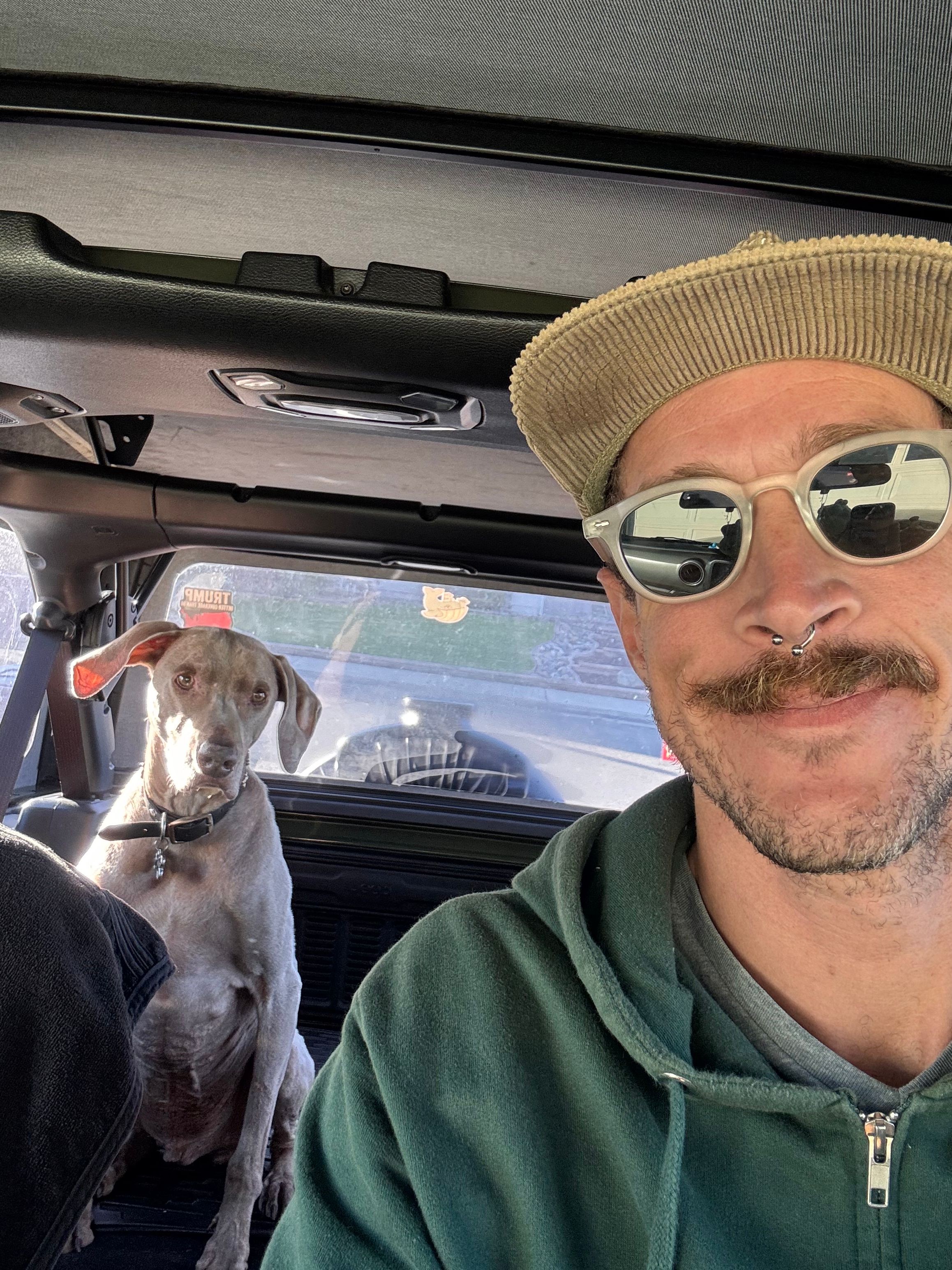 Man wearing sunglasses and a hat sitting in a vehicle with a dog beside him, both looking outside.