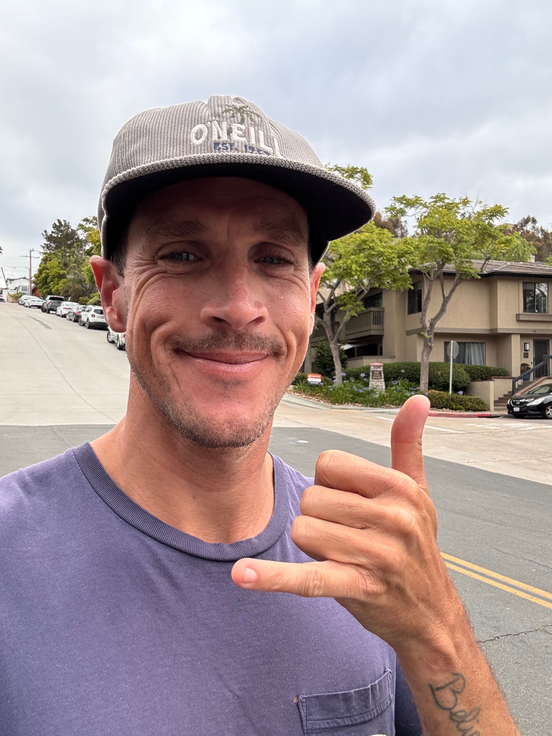 Man wearing a cap and purple shirt giving a thumbs-up outdoors, smiling, with trees and buildings in background.