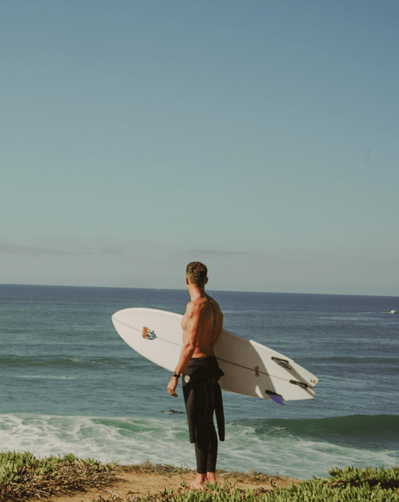 Person standing on grass holding a surfboard, facing the ocean with waves, under a clear sky.