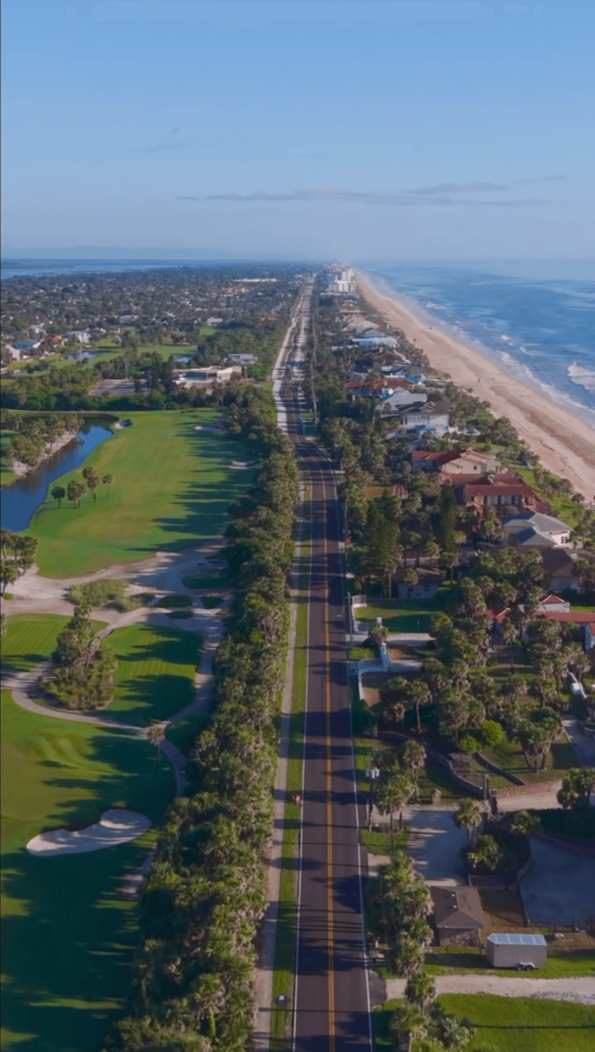 Aerial view of a coastal area with a road running parallel to the beach, green park, and residential buildings.