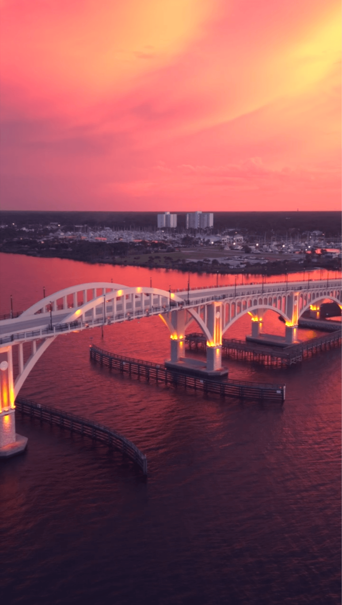 Bridge over water with city skyline and colorful sunset sky in background.