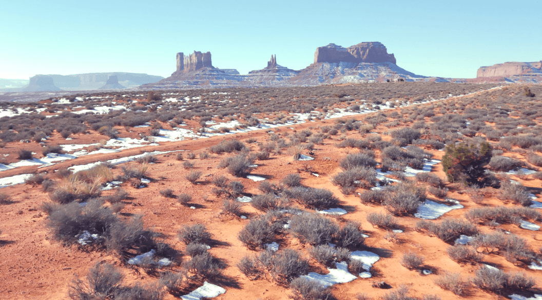 Desert landscape with sparse vegetation and snow patches, with mesas and buttes in the background under a clear blue sky.