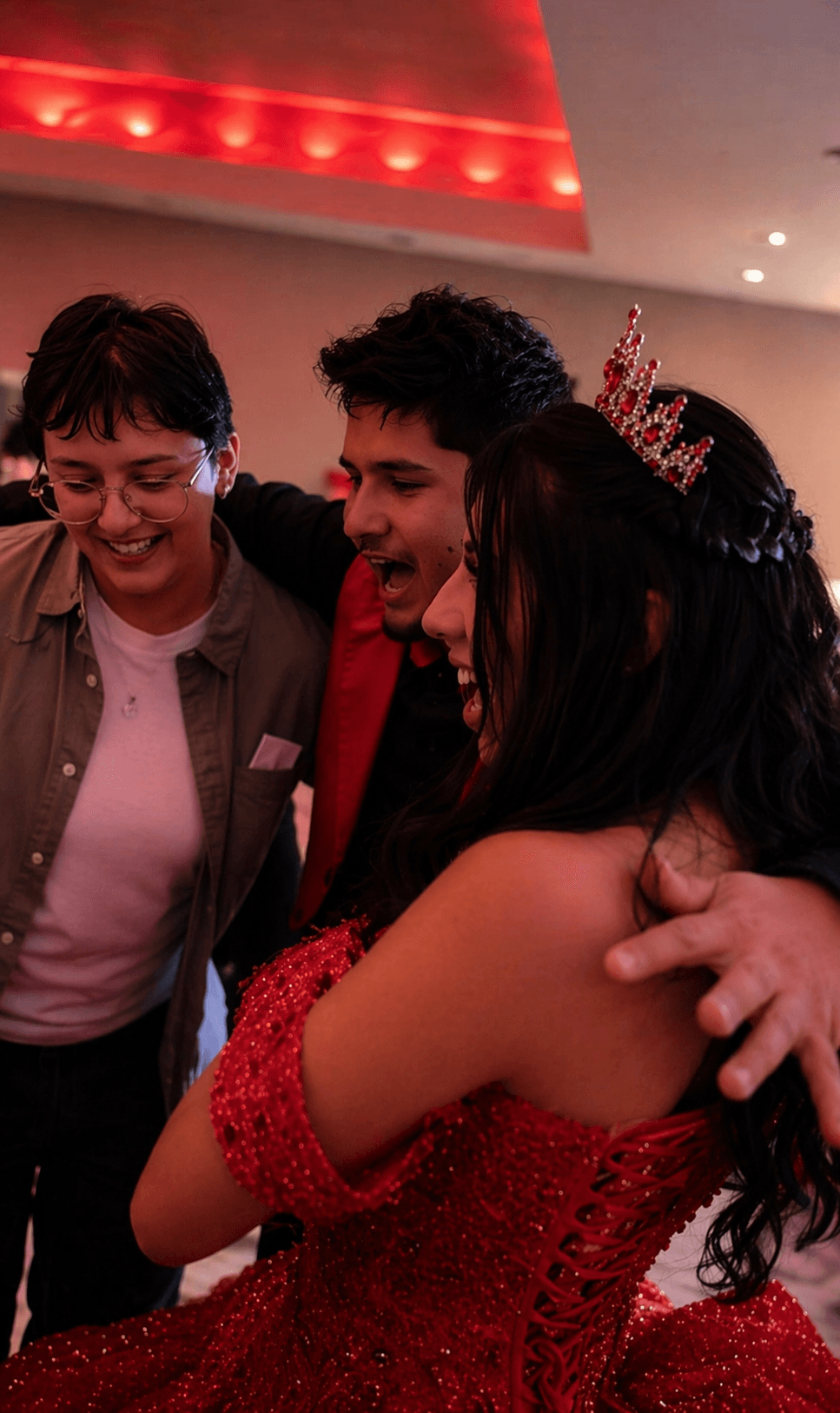 Three people smiling and hugging, one wearing a tiara and red dress, in a decorated indoor setting.