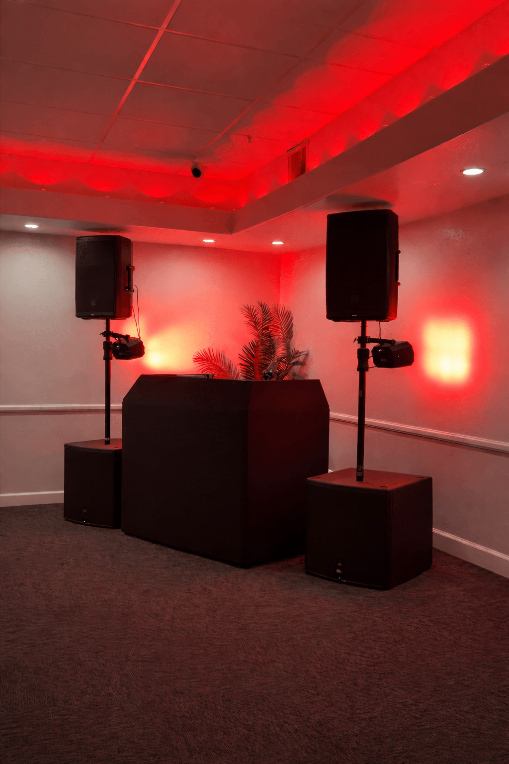Setup with two large speakers on stands, a black sofa, and red lighting on the wall, in a room with carpeted floor.