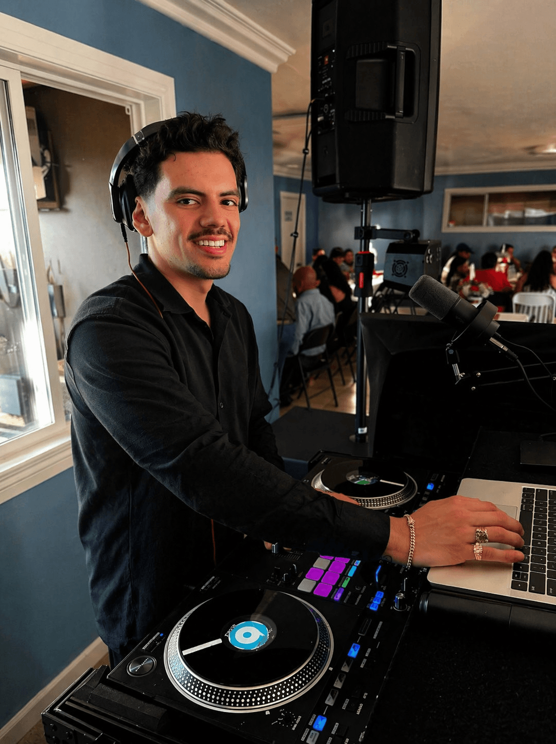 DJ smiling at turntables and laptop in a room with people in the background.