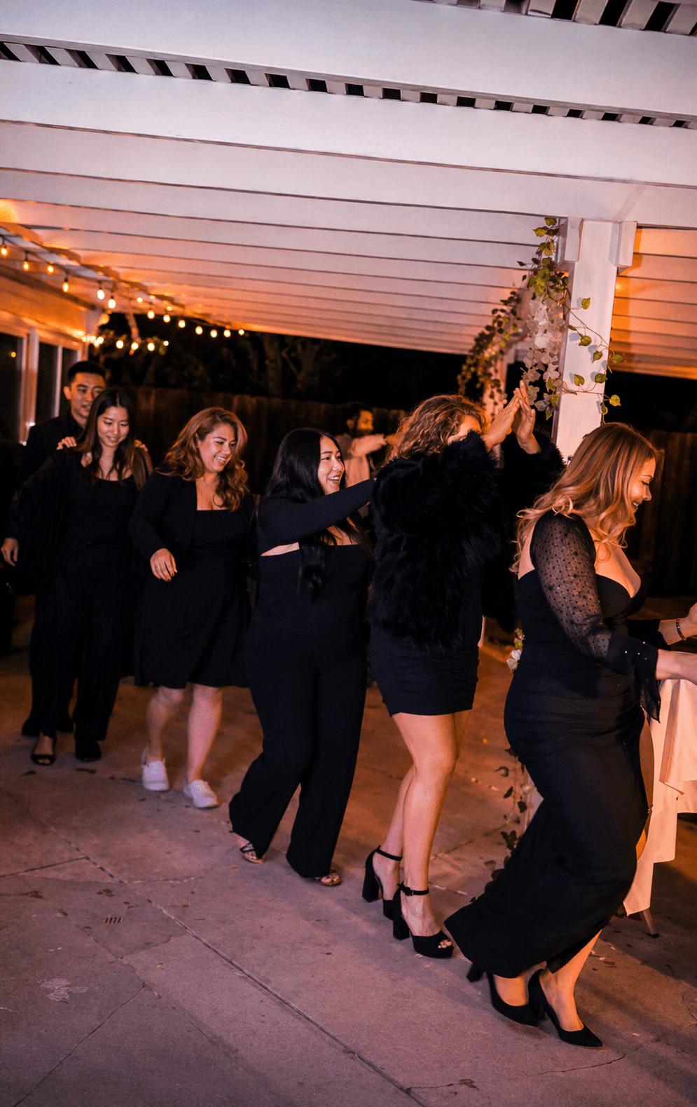 Group of women in black dresses standing in line under a decorated outdoor ceiling with string lights, at night.
