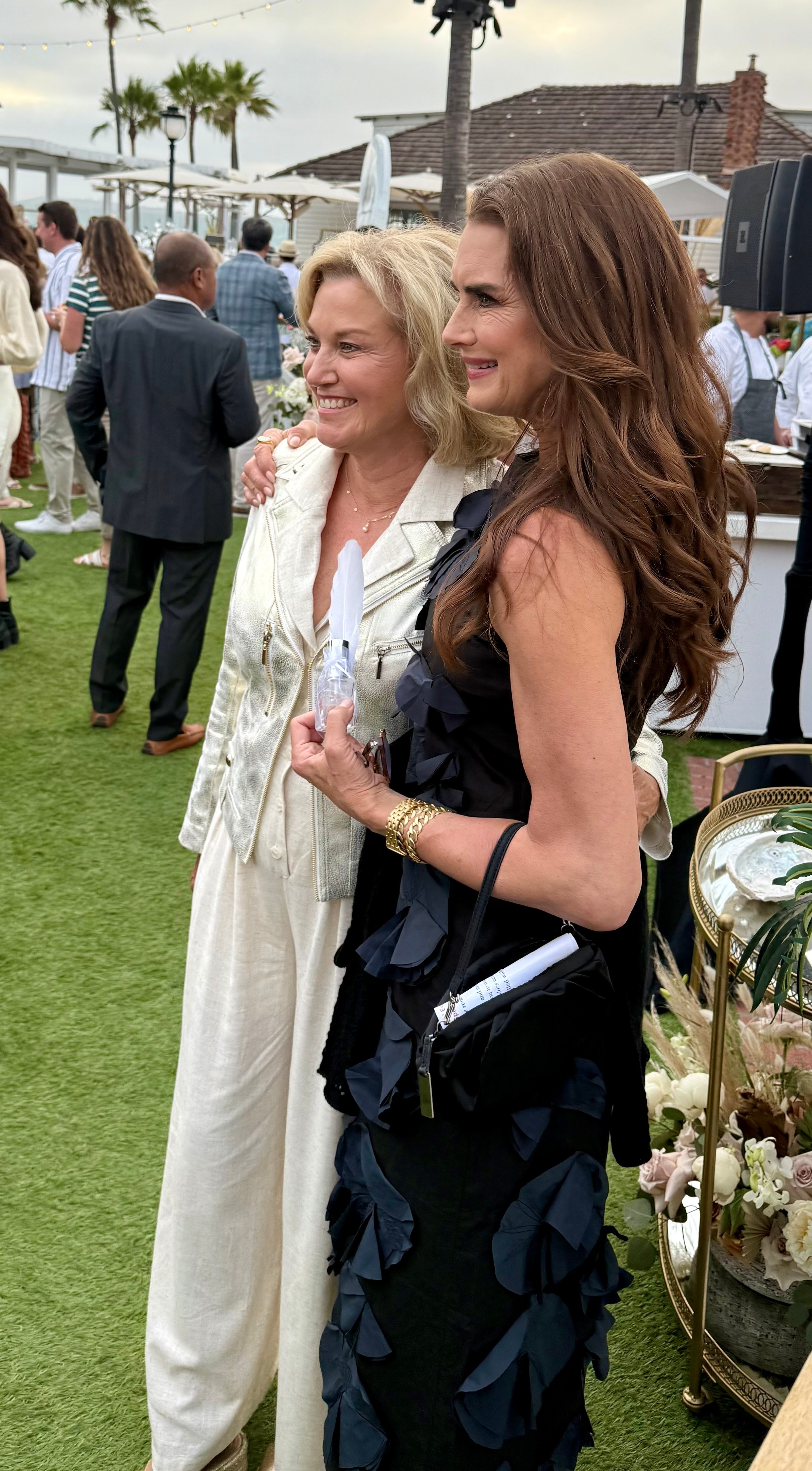 Two women smiling and posing outdoors at an event, with people and palm trees in the background.