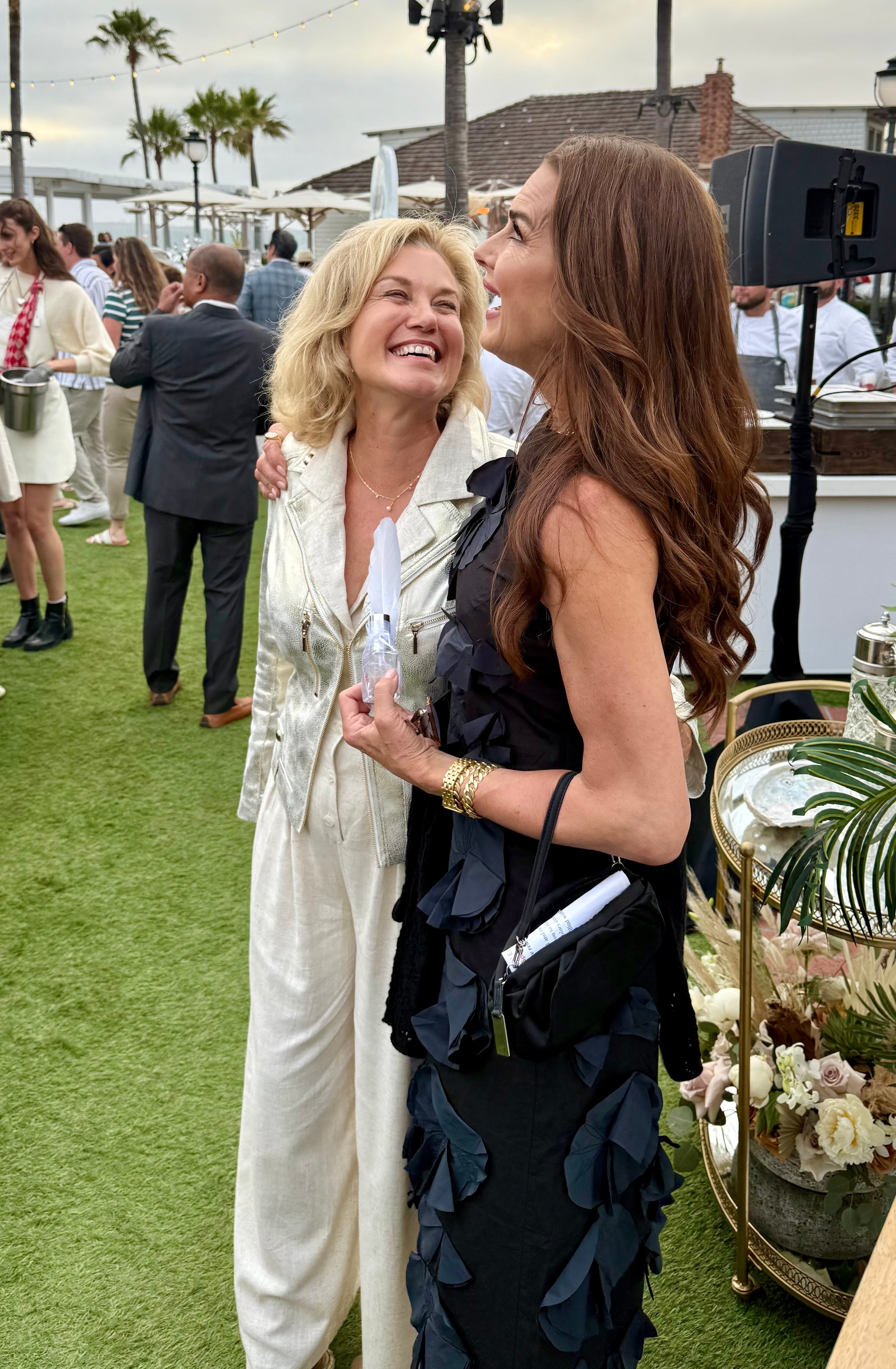 Two women smiling and talking outdoors at an event, with people and palm trees in the background.