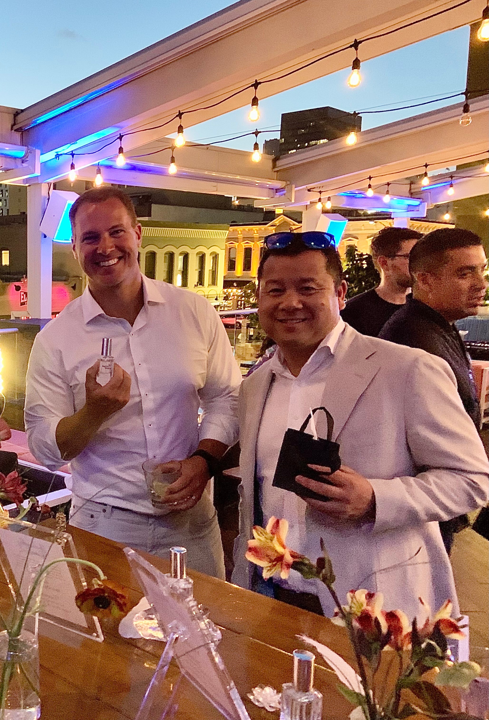 Two men smiling at a social event, one holding a drink and the other holding a phone, under string lights.