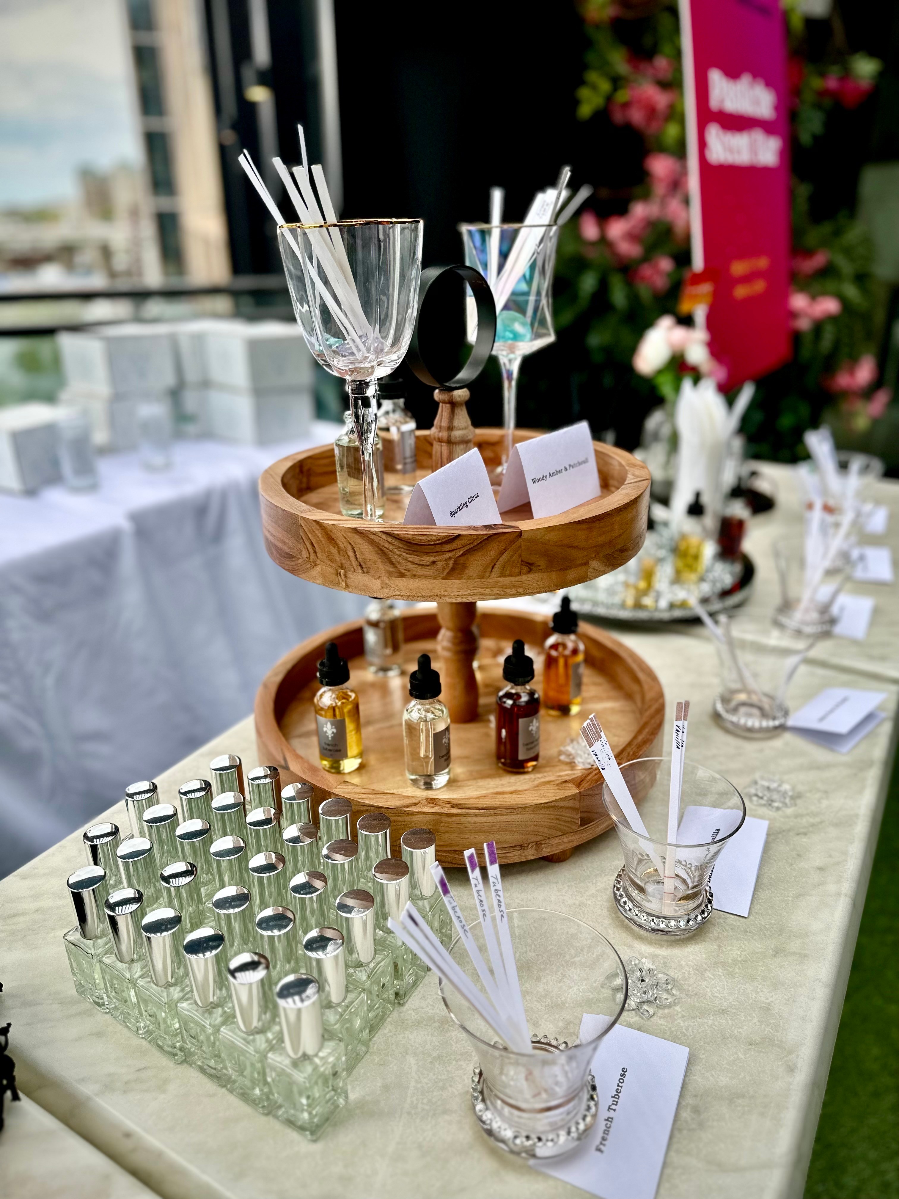 Table with glassware, bottles, and a wooden stand, set outdoors with flowers and a pink sign in the background.