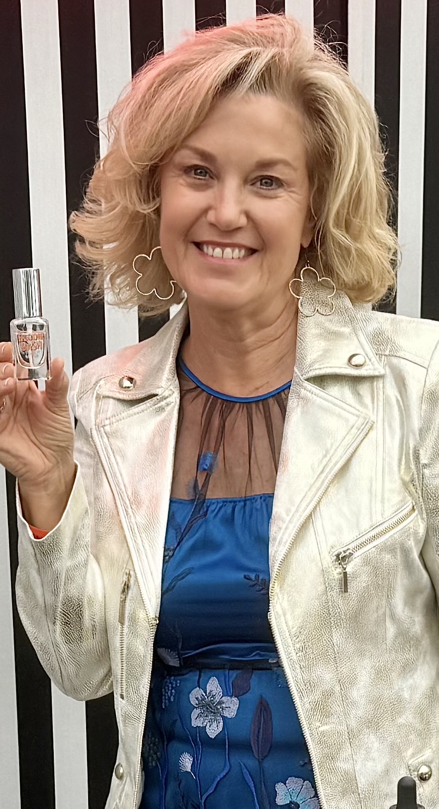 Woman with blonde hair smiling, holding a small bottle, wearing a white jacket and blue dress, standing in front of black and white striped background.