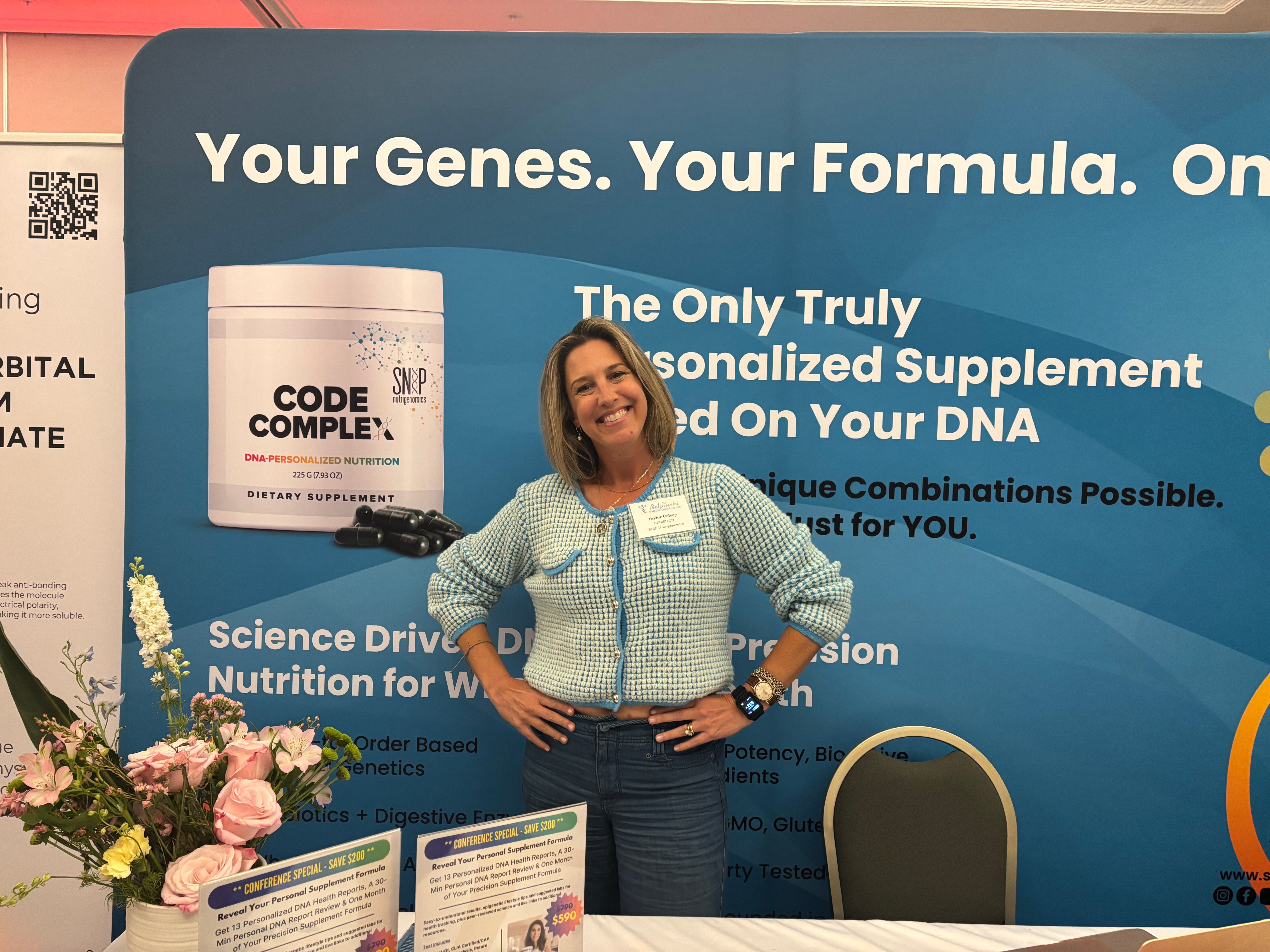 Woman standing in front of a blue promotional backdrop, smiling with hands on hips, flowers on the left, and a chair on the right.