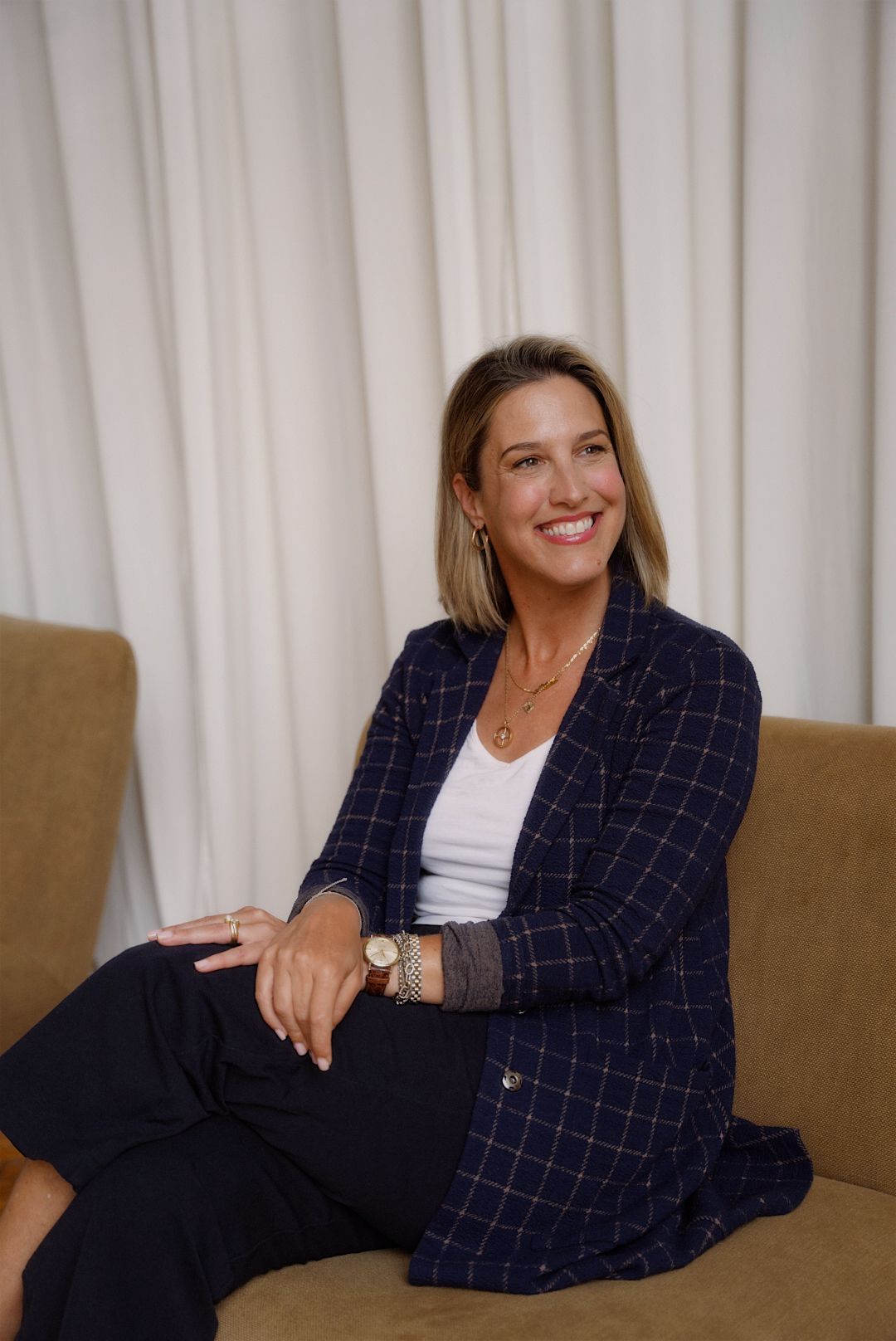 Woman sitting on a couch, smiling, with a white curtain background, wearing a dark blazer and checkered pants.