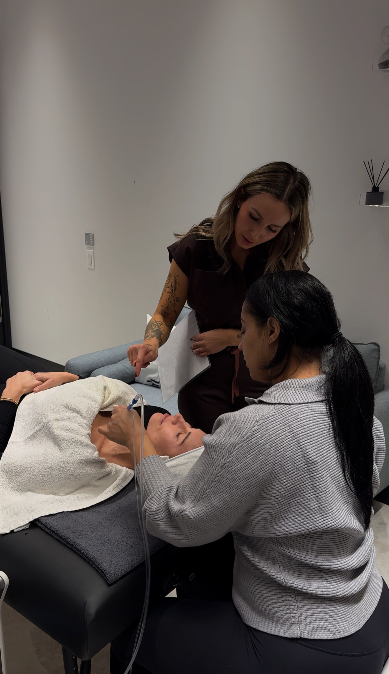 Two women are attending to a person lying on a medical bed, with one woman standing and the other sitting nearby.