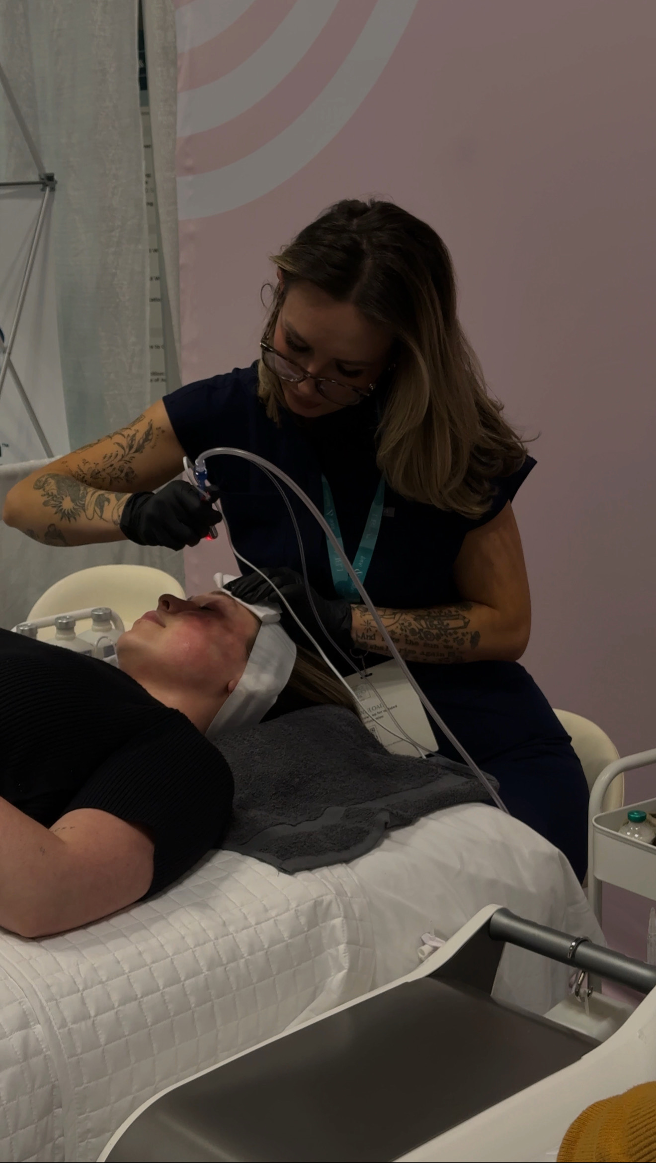 A woman with glasses and tattoos performs a medical procedure on a patient lying in bed, with medical equipment nearby.