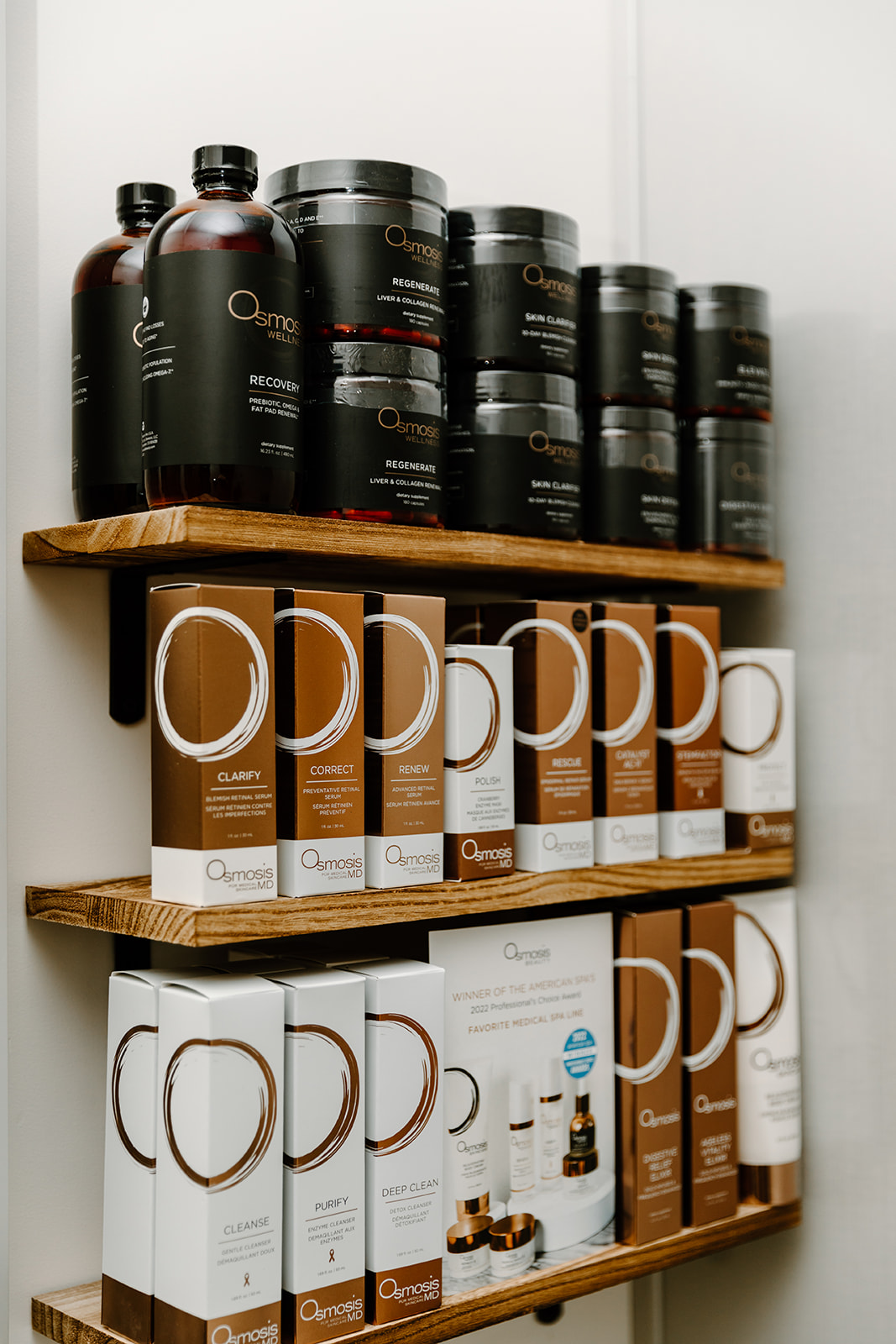 Three wooden shelves hold black and white bottles and boxes with product labels, arranged vertically against a white wall.