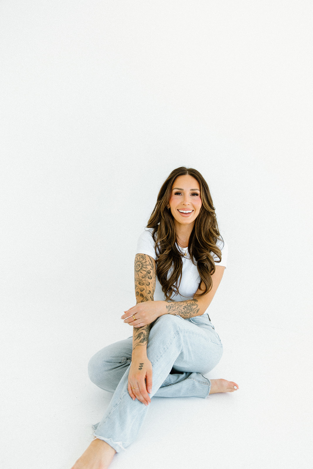 Young woman with long hair, smiling, sitting cross-legged on the floor against a plain white background.