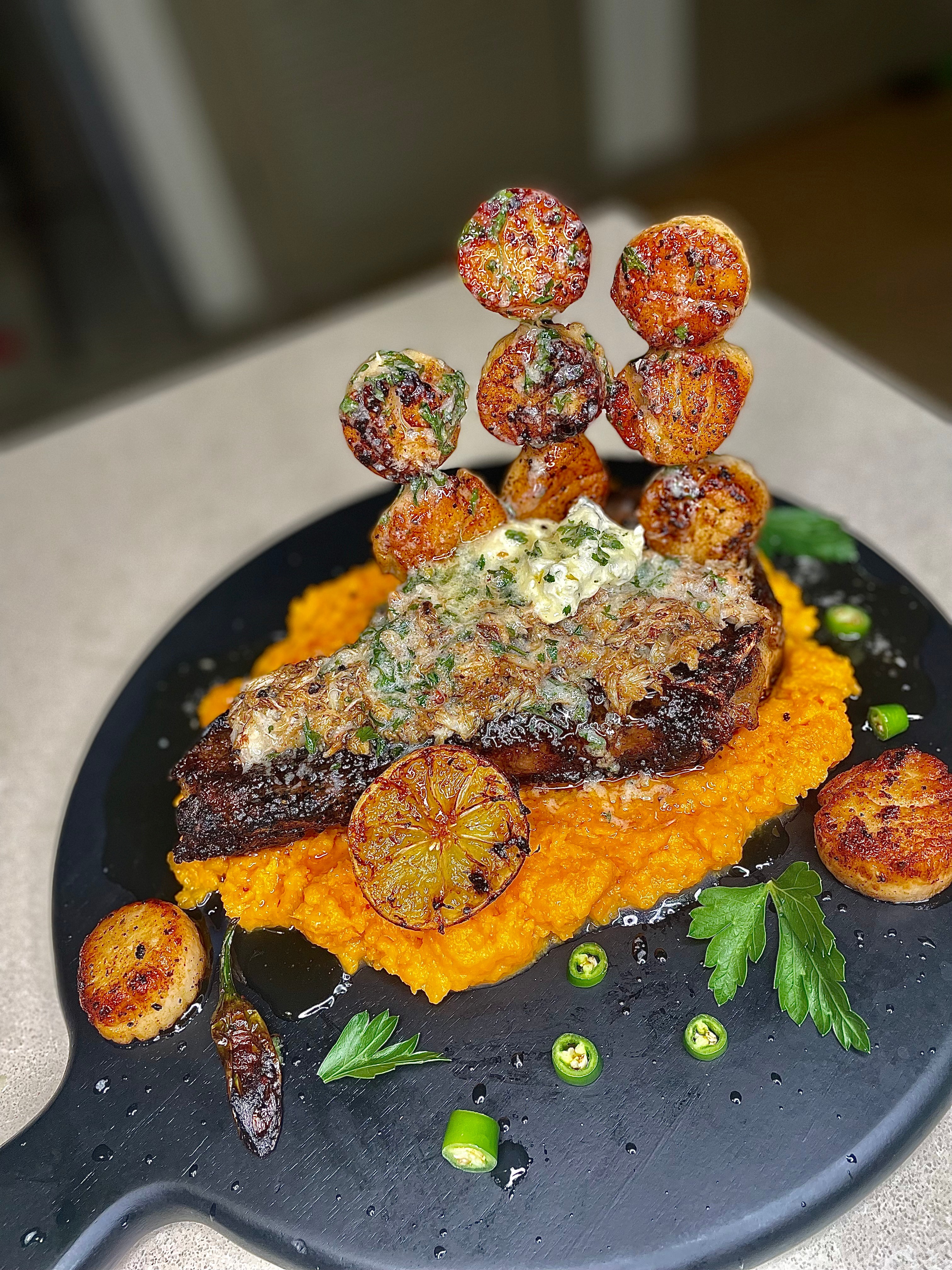 A plated dish with a piece of meat topped with herbs, surrounded by roasted vegetables, green onions, and garnishes on a black plate.