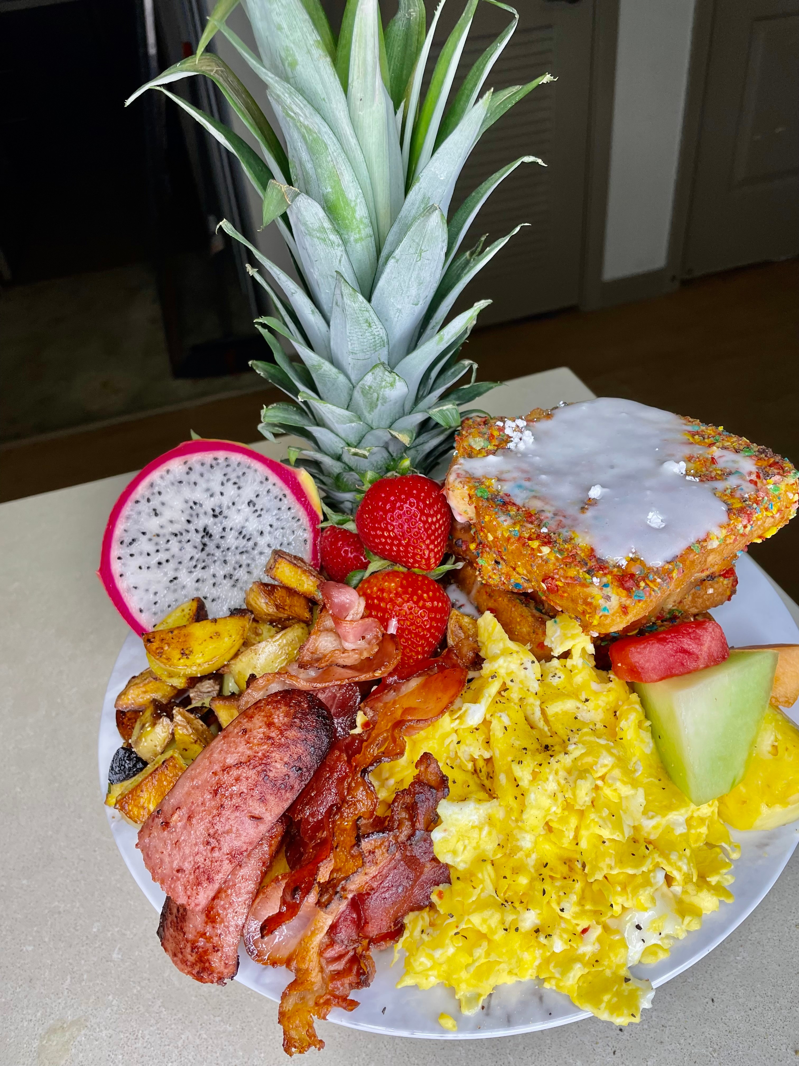 Pineapple, strawberries, scrambled eggs, bacon, sausage, toast with icing, fruit slices, and a dragon fruit half on a plate.