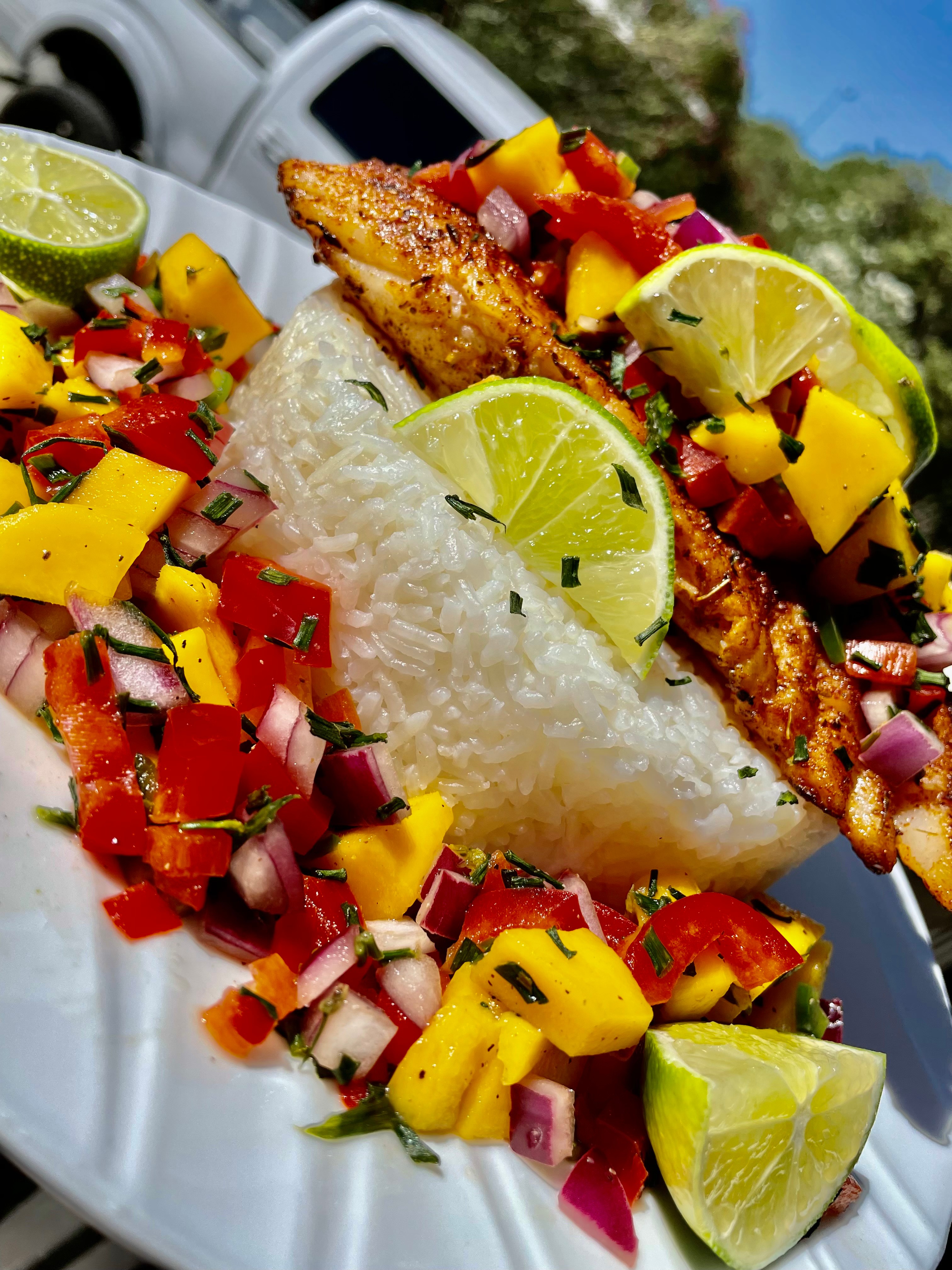 Plate with grilled fish, rice, and colorful mango salsa with lime wedges, outdoors on a sunny day.