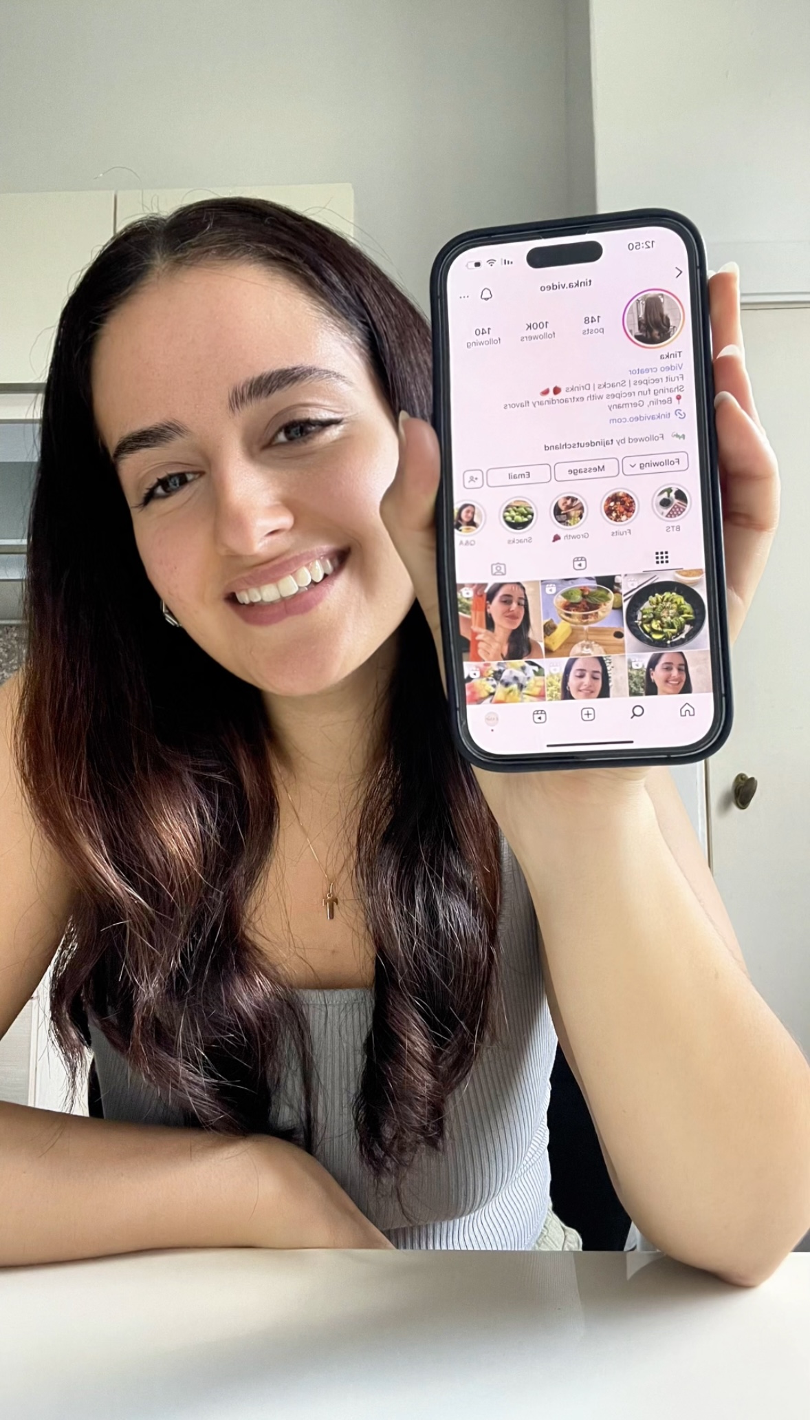 Young woman with long dark hair smiling, holding a smartphone displaying a social media profile, sitting at a table.