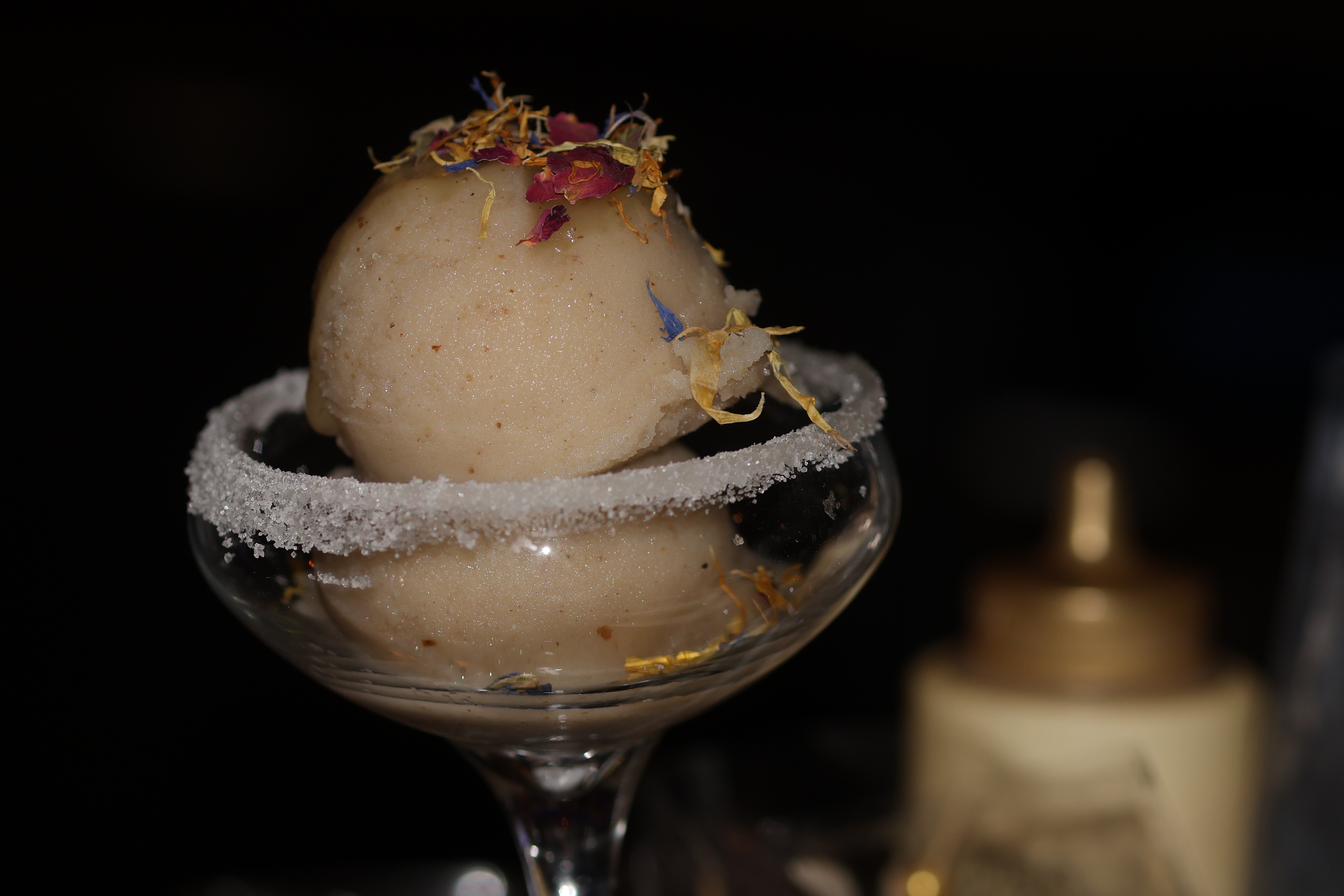 A scoop of ice cream in a glass dish with a salted rim, garnished with edible flowers, against a dark background.