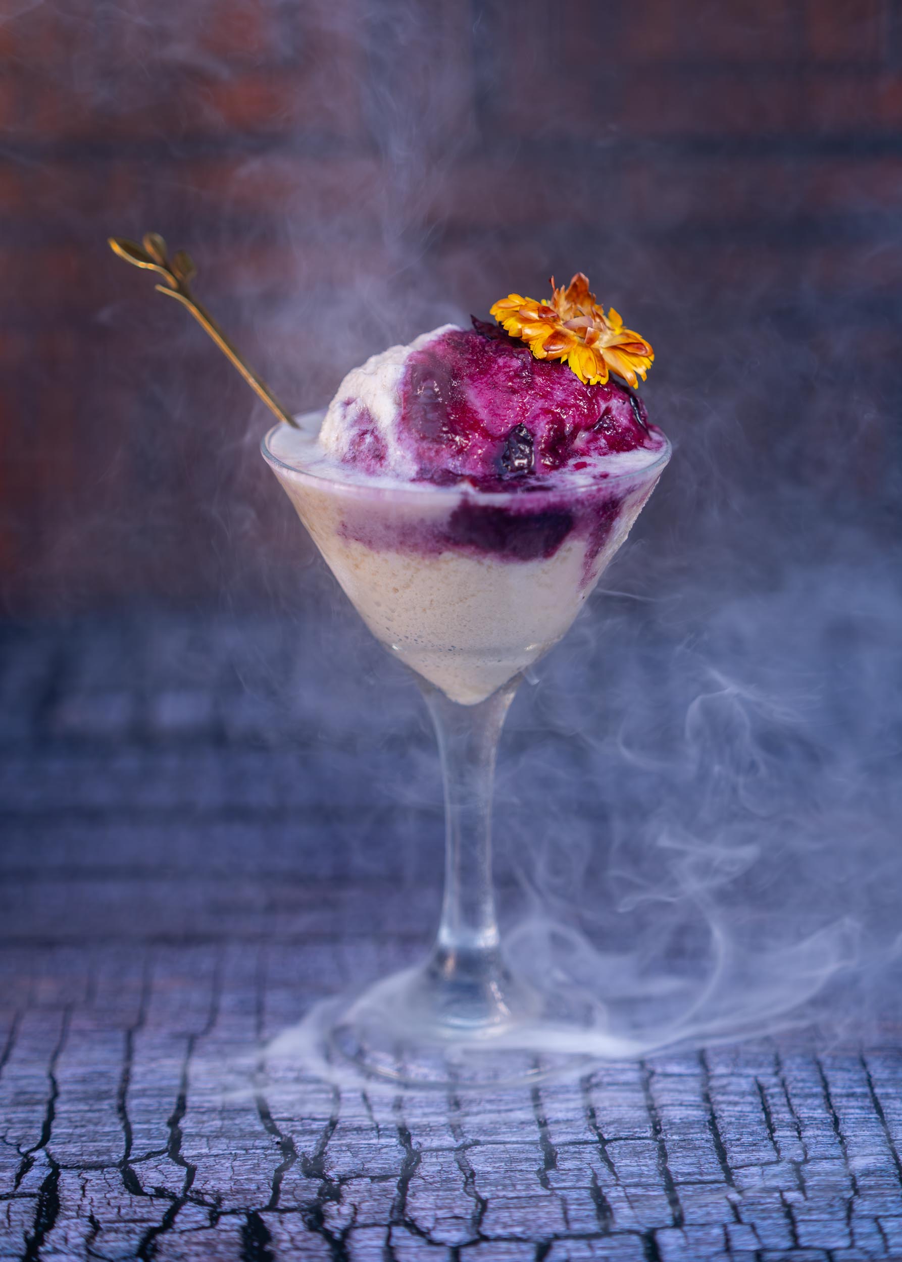 Dessert in a glass with purple and white layers, garnished with a flower and a decorative pick, surrounded by smoke.