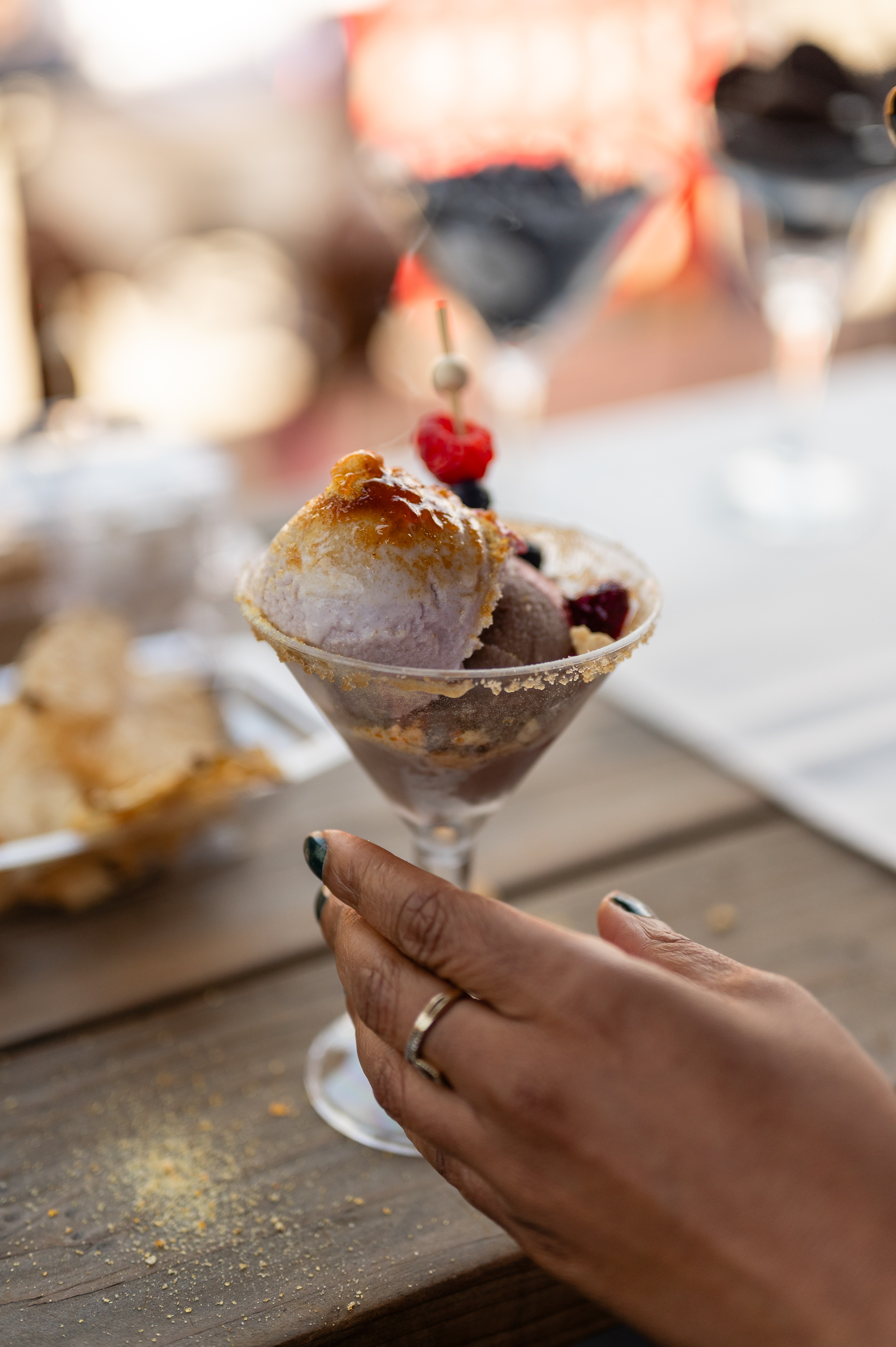 Person's hand holding a dessert glass with ice cream, berries, and a cherry on top, on a wooden table.