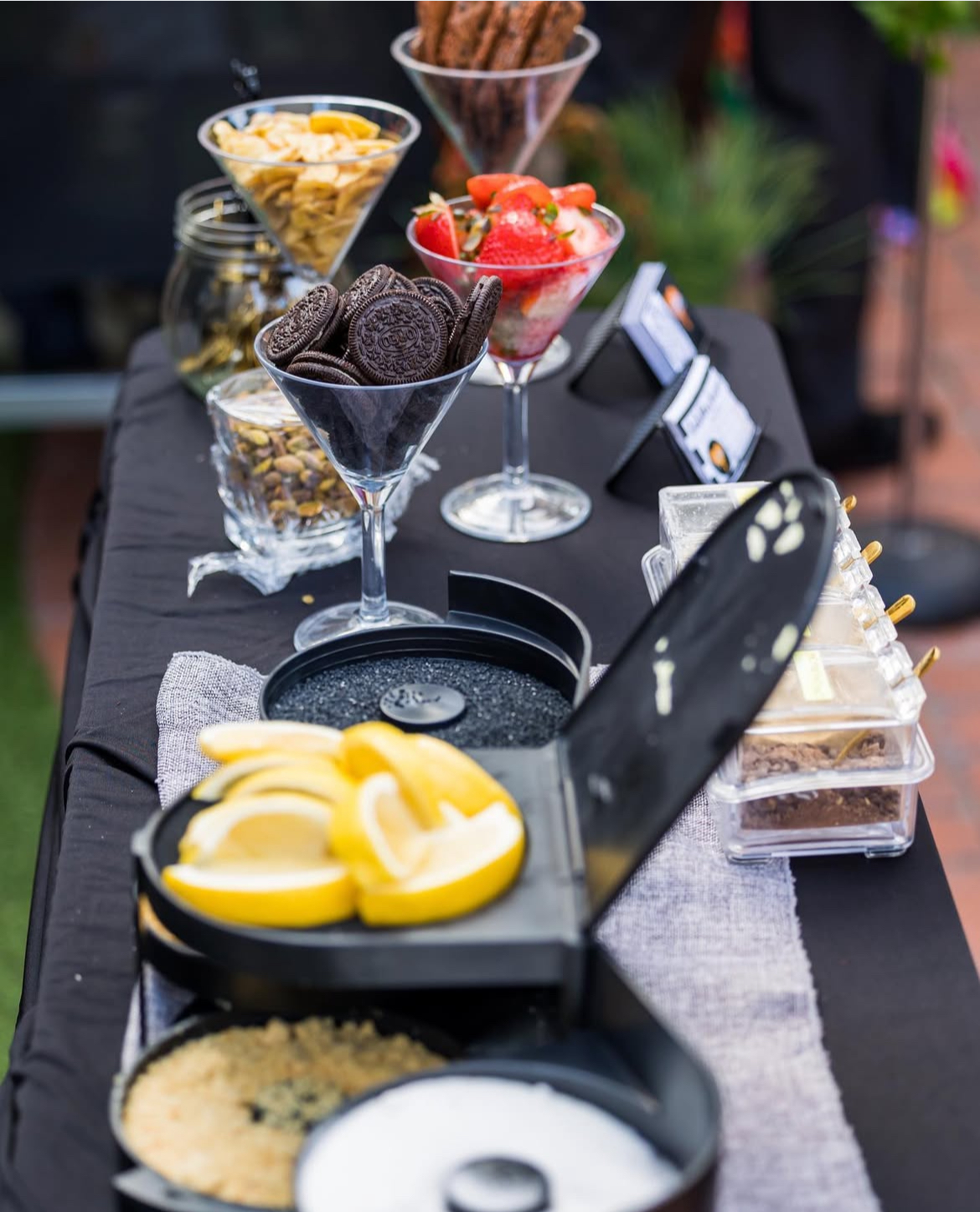 Assorted desserts and toppings in glass bowls and containers on a black table, with lemon slices and a black box in foreground.