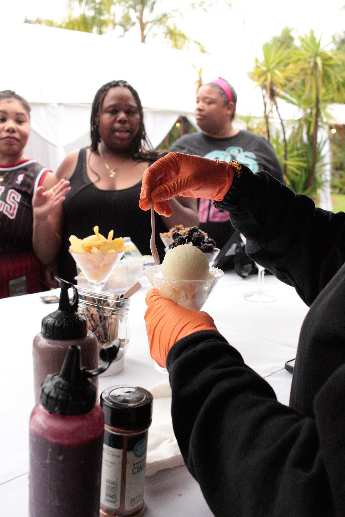 Person wearing orange gloves serving ice cream to three women outdoors with trees in background.