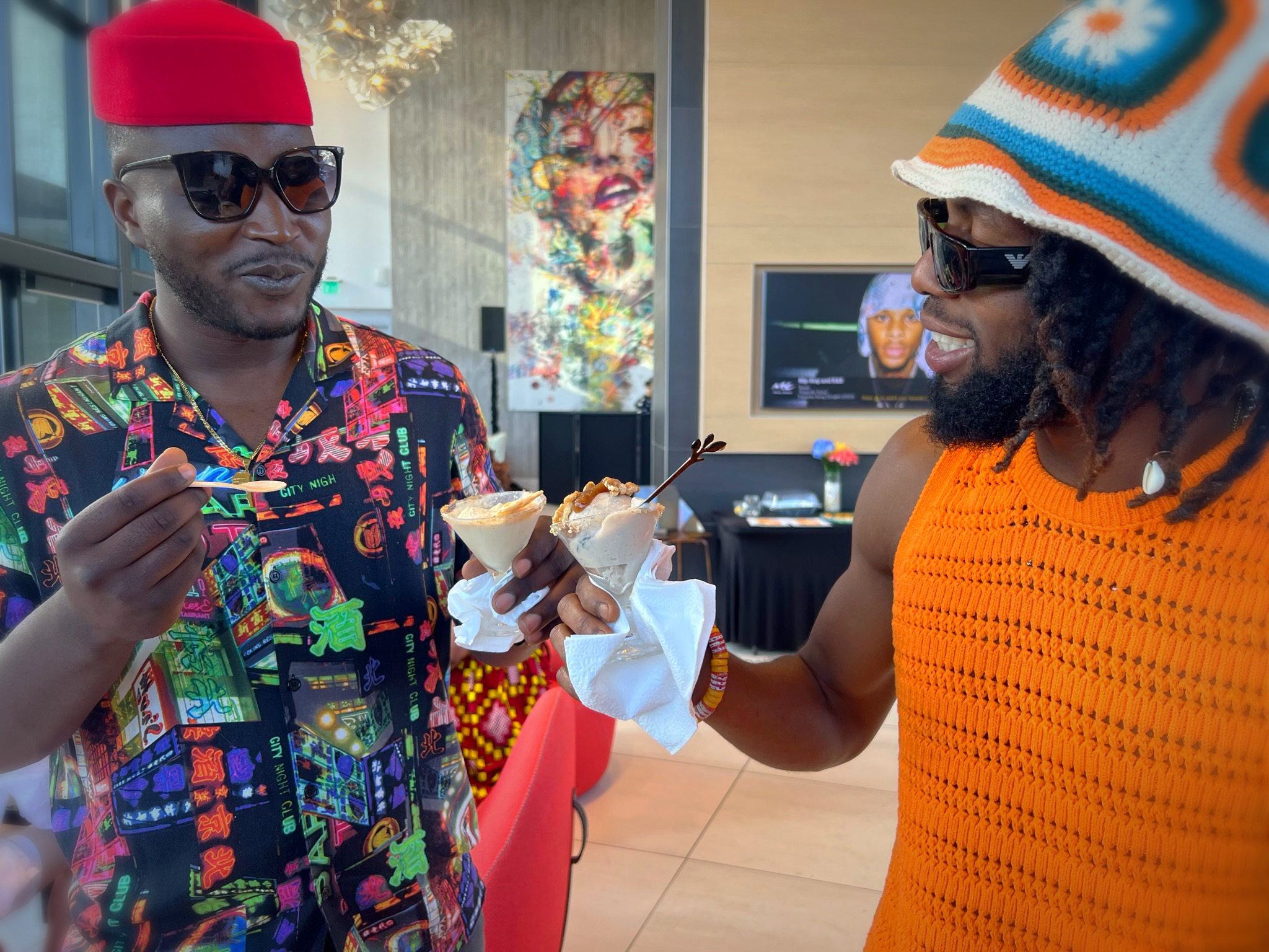 Two men with colorful clothing and accessories share a dessert in a room with a TV and artwork.