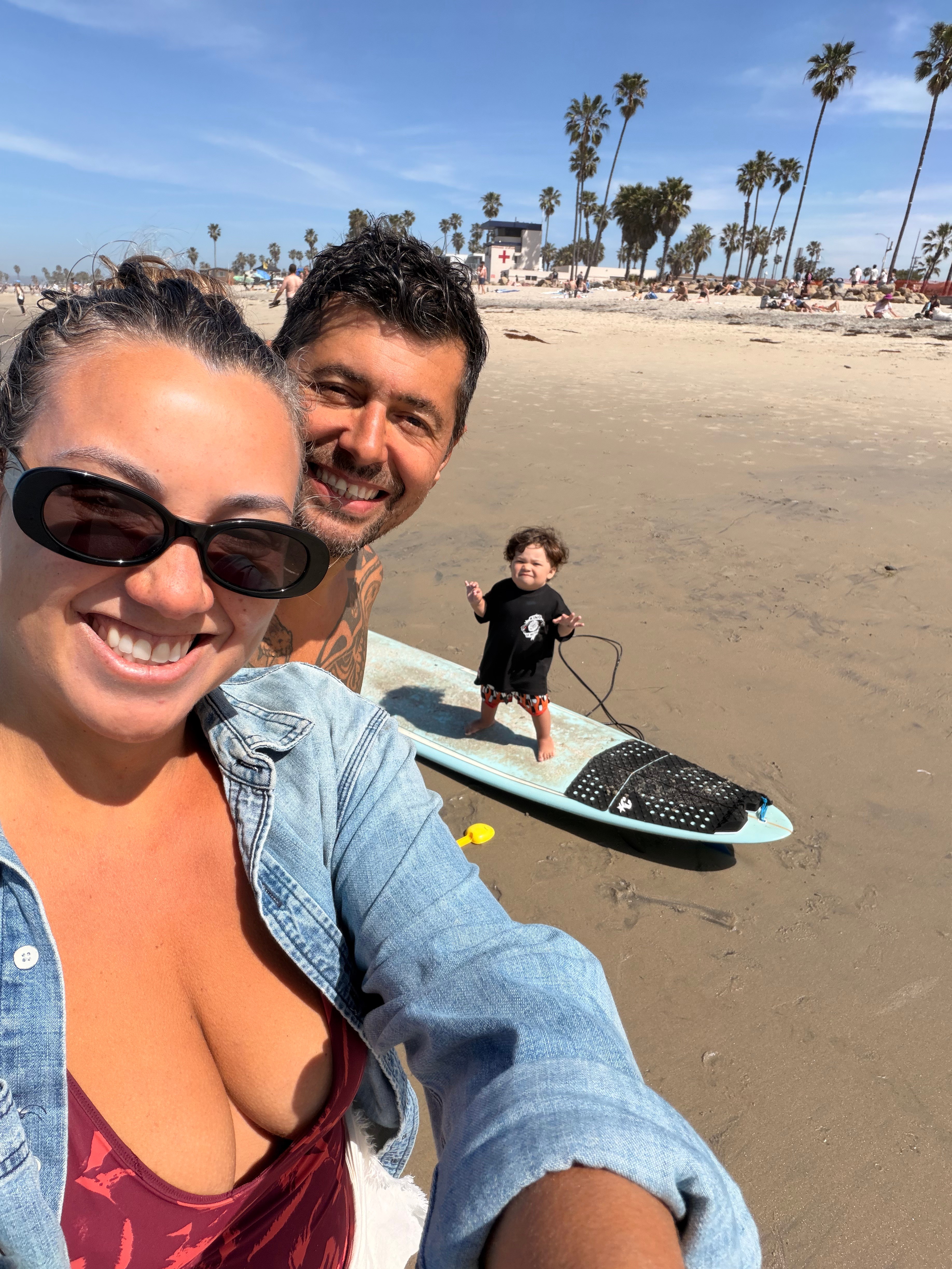 Two adults and a child on a beach with a paddleboard, palm trees, and a clear blue sky in the background.