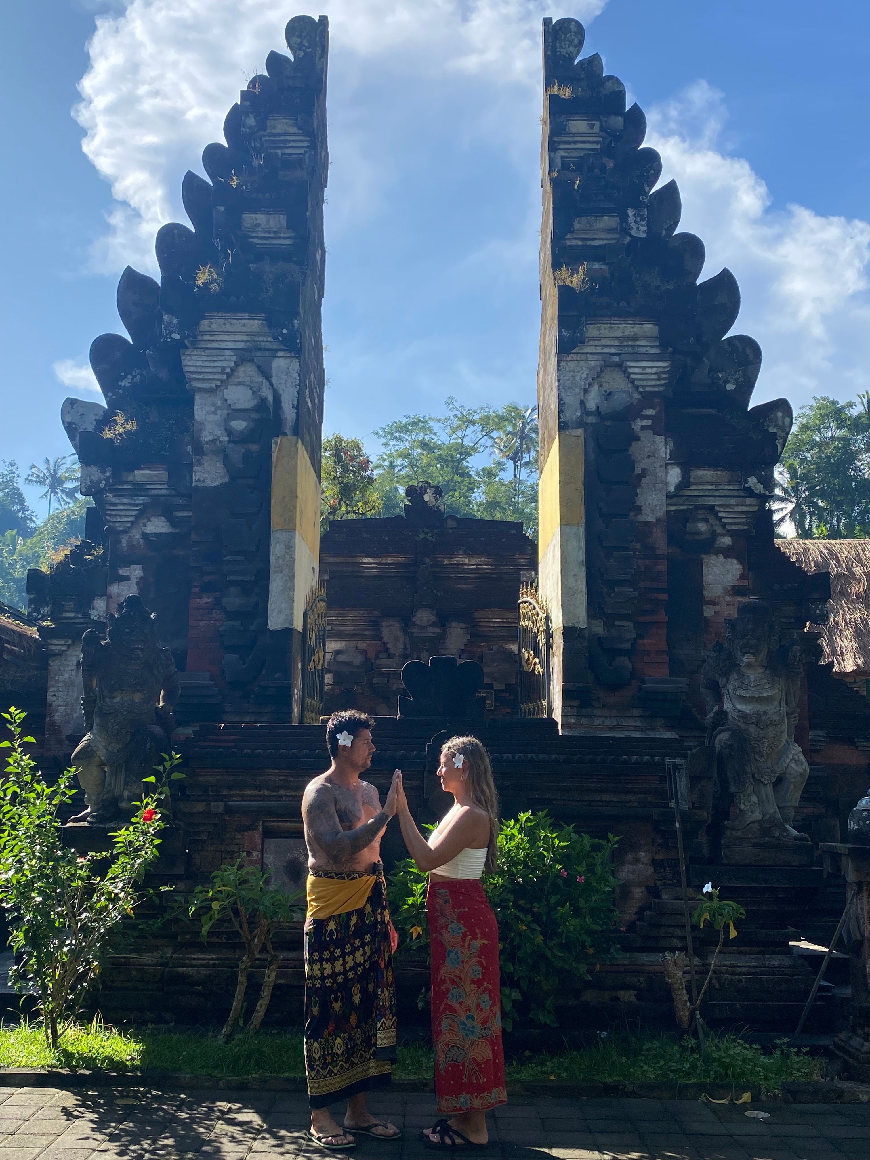 Two people stand near an ancient stone structure with a tiered, pointed top, surrounded by greenery and a blue sky.
