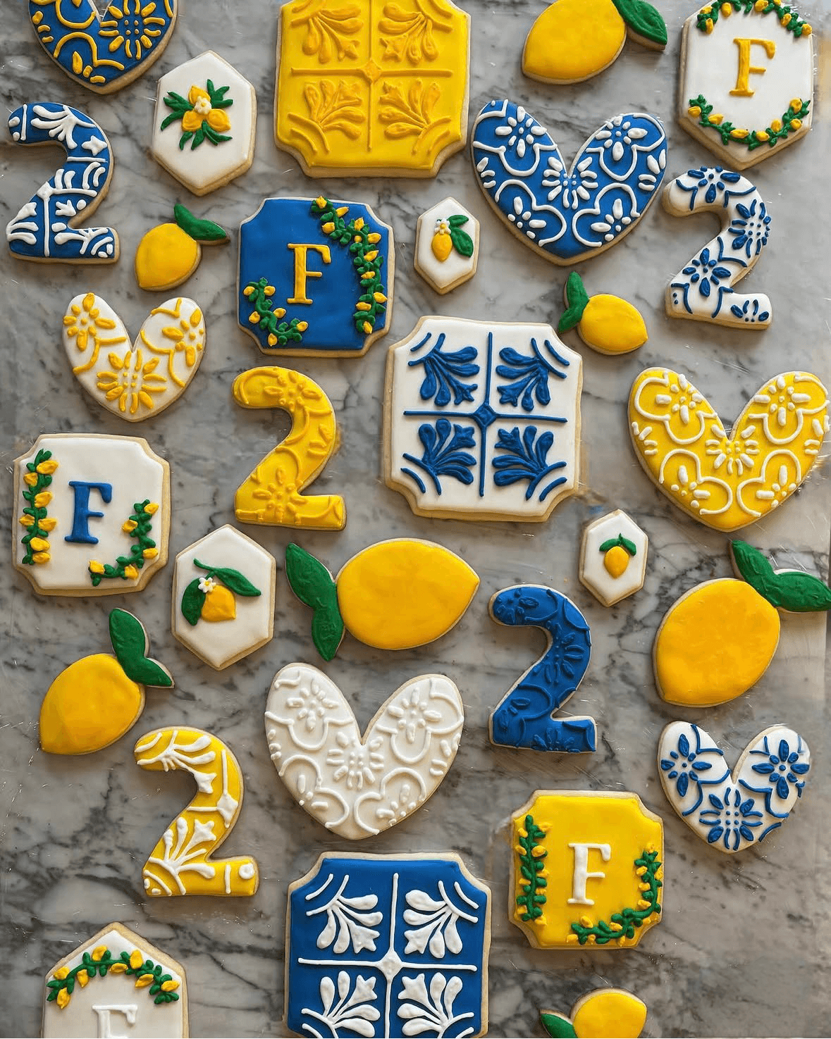 Decorative cookies in yellow, blue, and white with floral, heart, and letter designs, arranged on a gray surface.