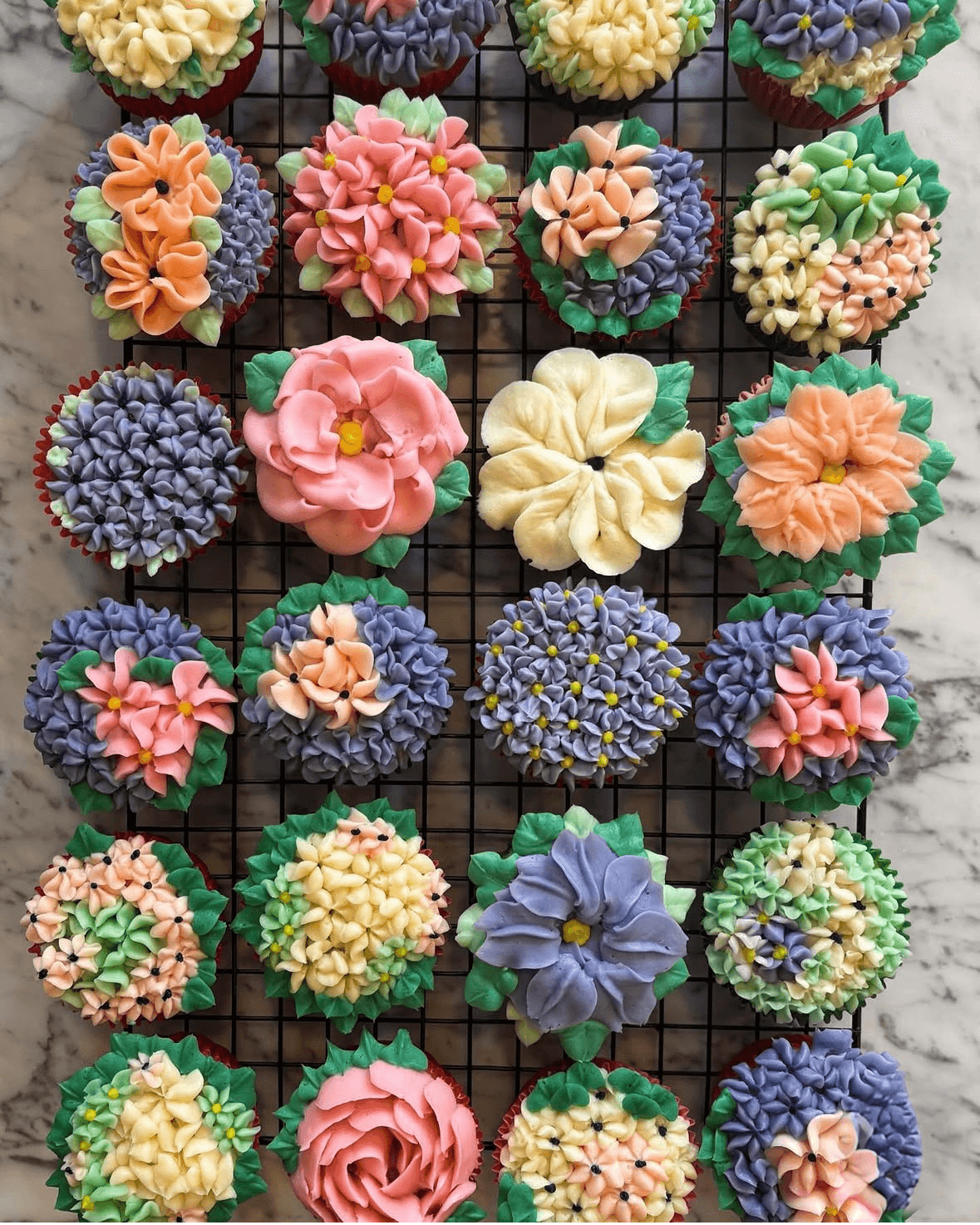 Assorted colorful cupcakes with floral and decorative frosting on a black wire rack.