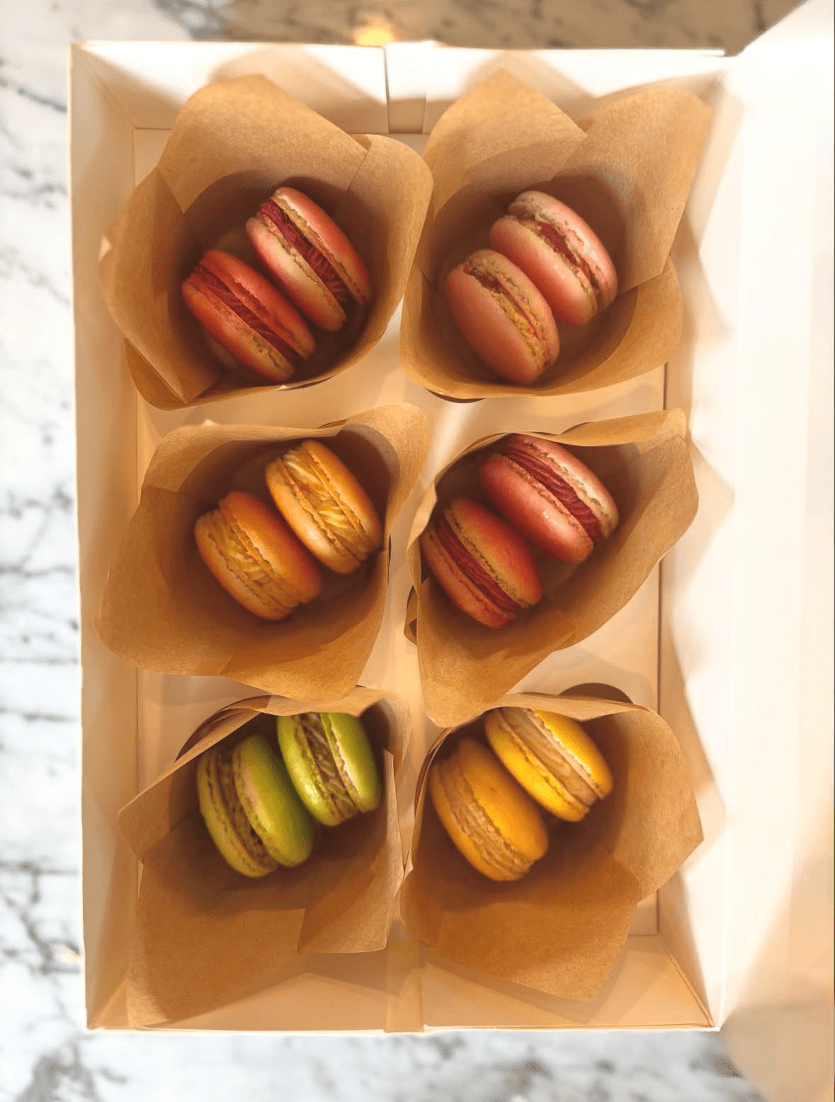 Six colorful macarons in a box, arranged in two rows of three, with brown paper wrappers, in various colors including pink, yellow, and green.