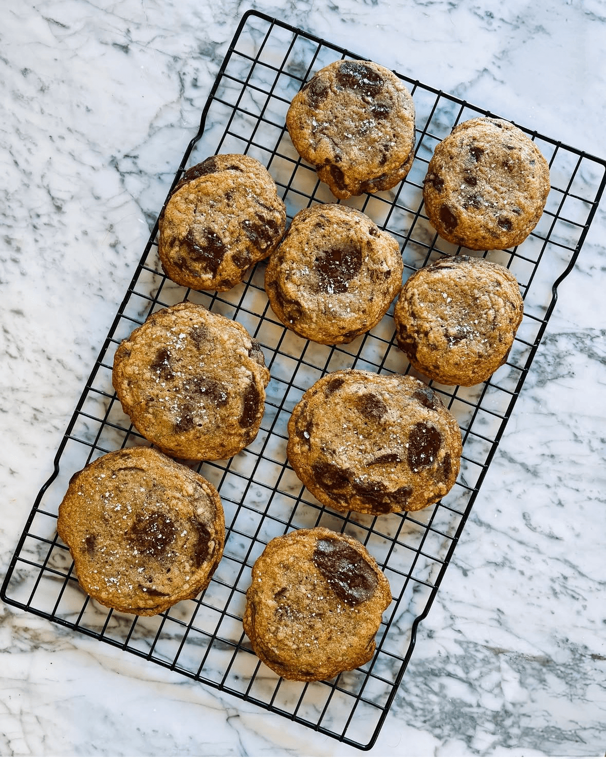 Ten chocolate chip cookies on a wire rack on a marble surface, some with visible chocolate chips.